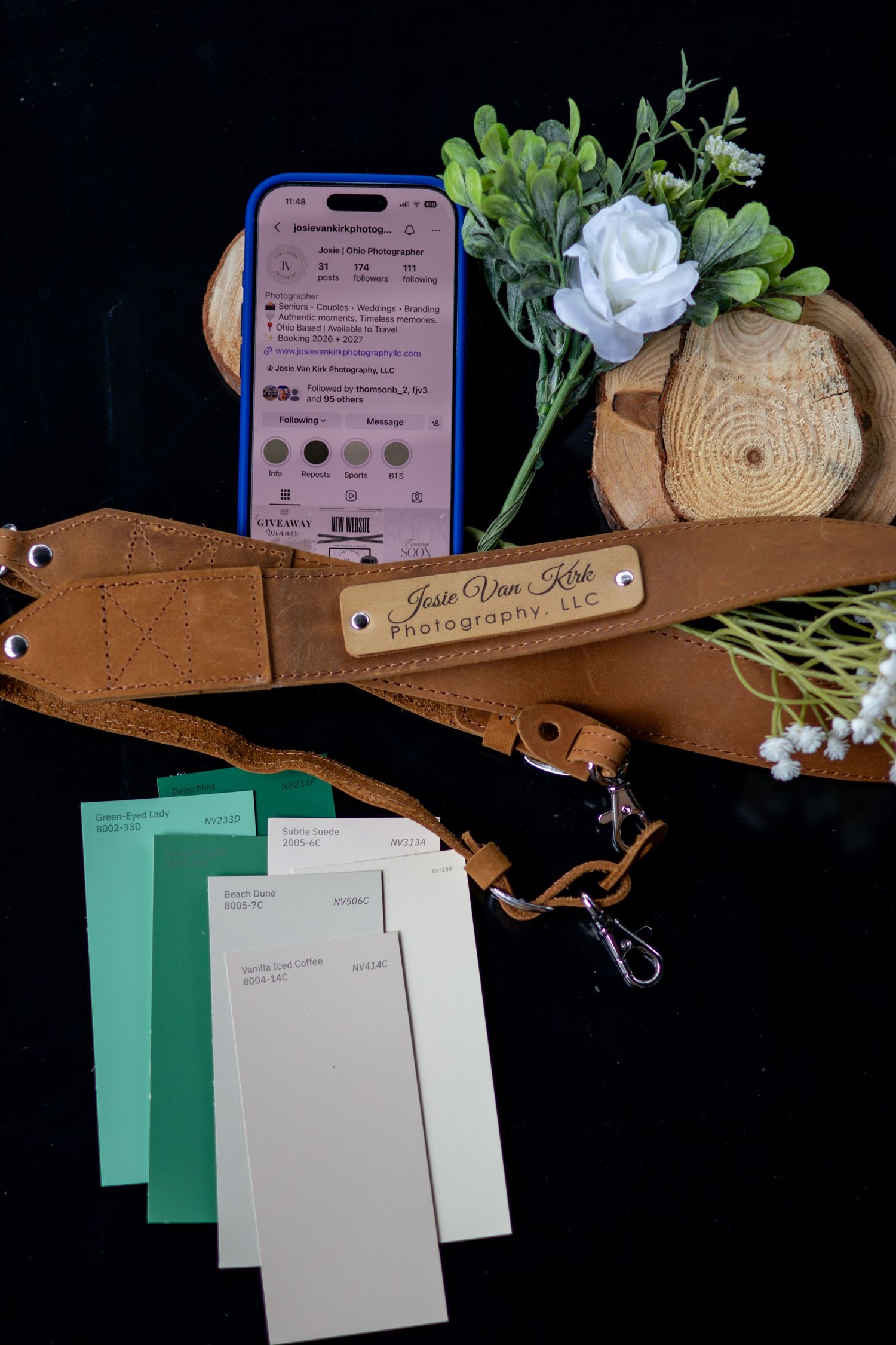 Camera strap engraved with Jessie Van Kirk Photography LLC beside paint swatches, phone, and white flowers on black background.
