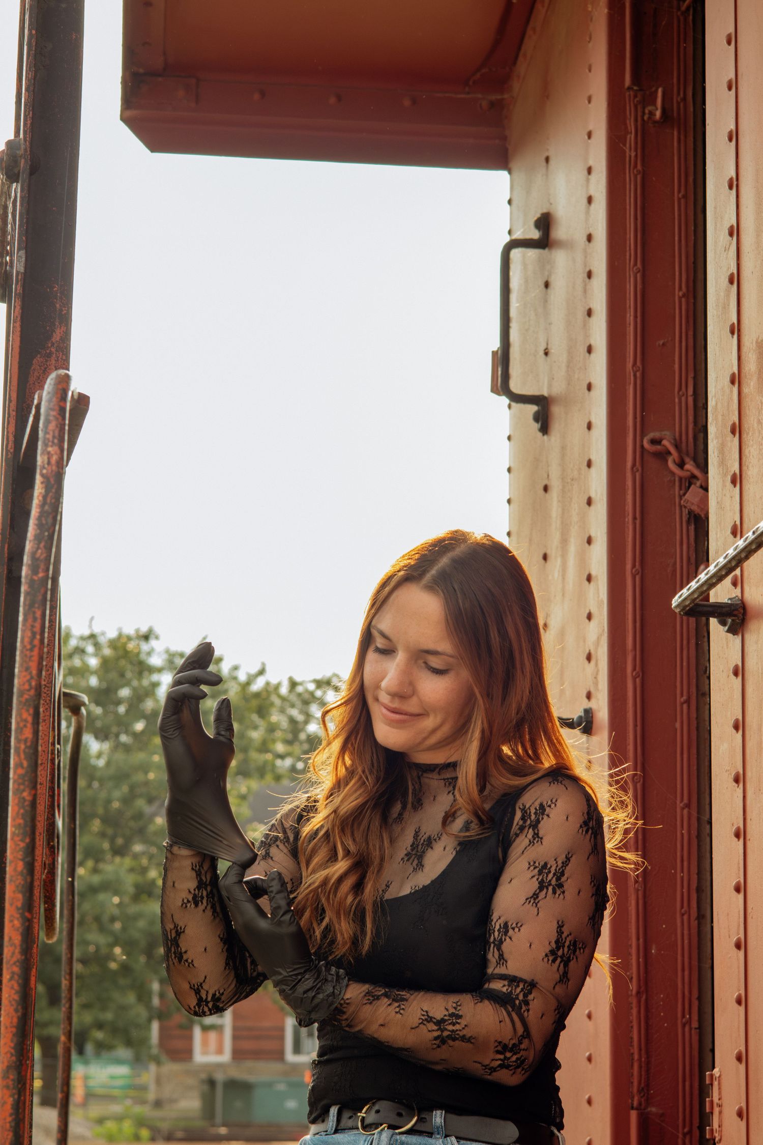 Young woman in black lace top standing in doorway of a vintage red train car at golden hour.