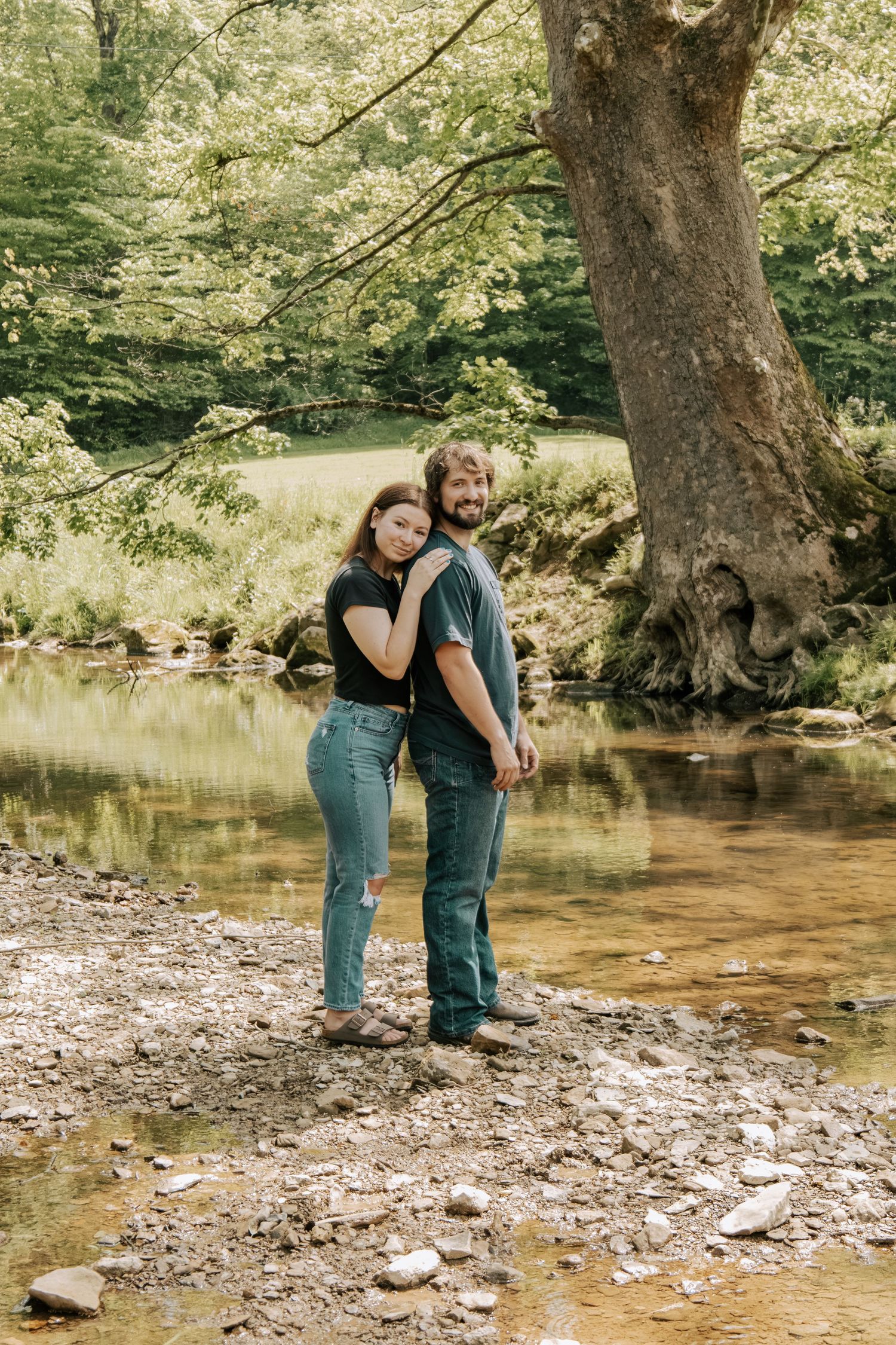Happy couple posing together by a rocky creek with a large tree and green forest backdrop.