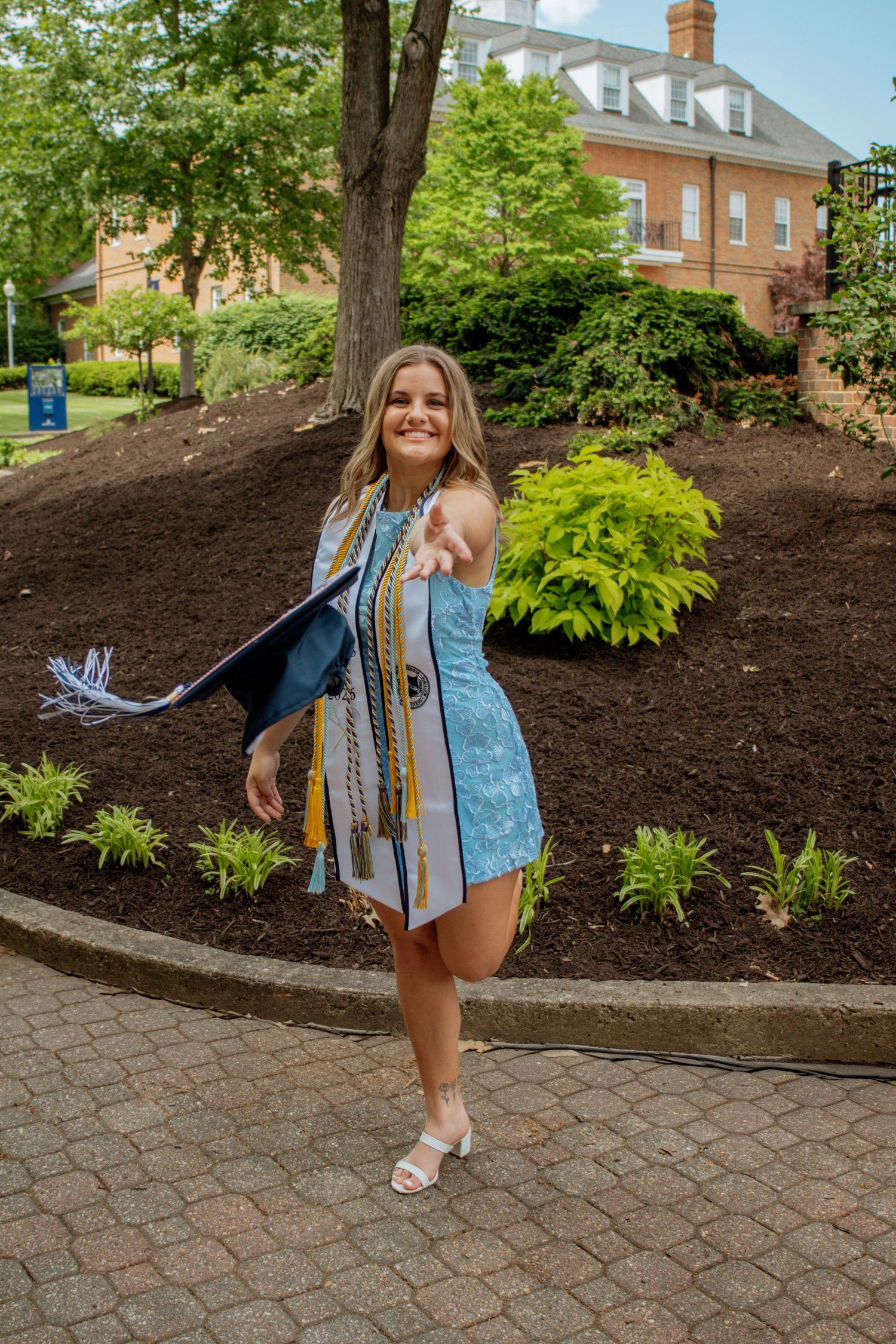 Smiling female graduate in blue dress holding cap and diploma on college campus brick pathway.