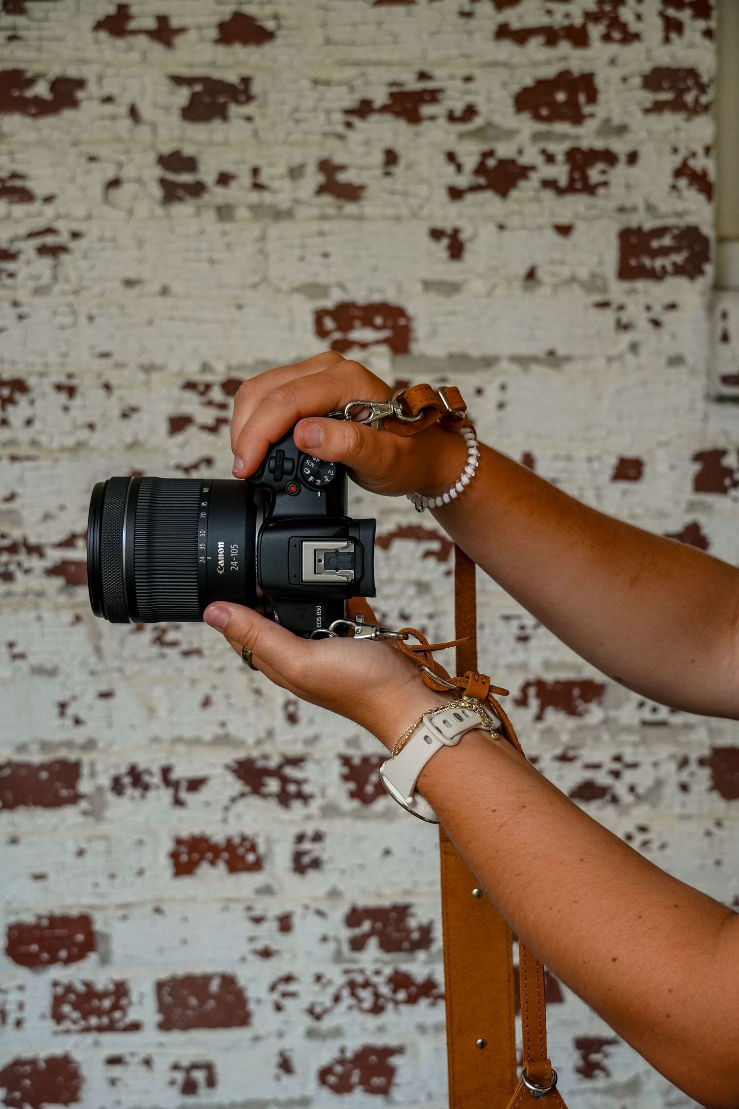 Photographer adjusting Canon camera with branded leather strap against rustic white brick wall.