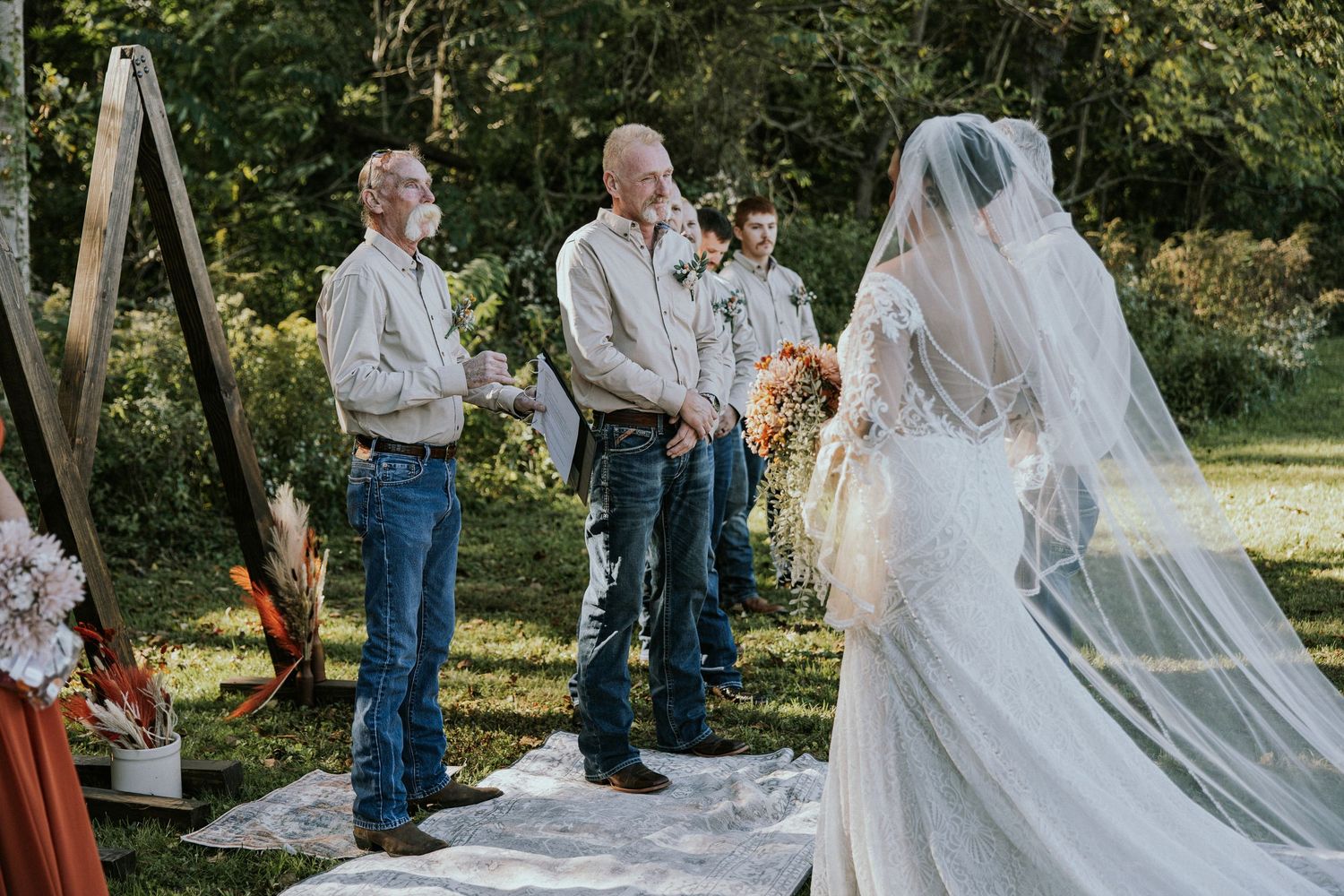 Outdoor wedding ceremony with bride in lace gown and veil approaching groom and officiant on a rustic altar.