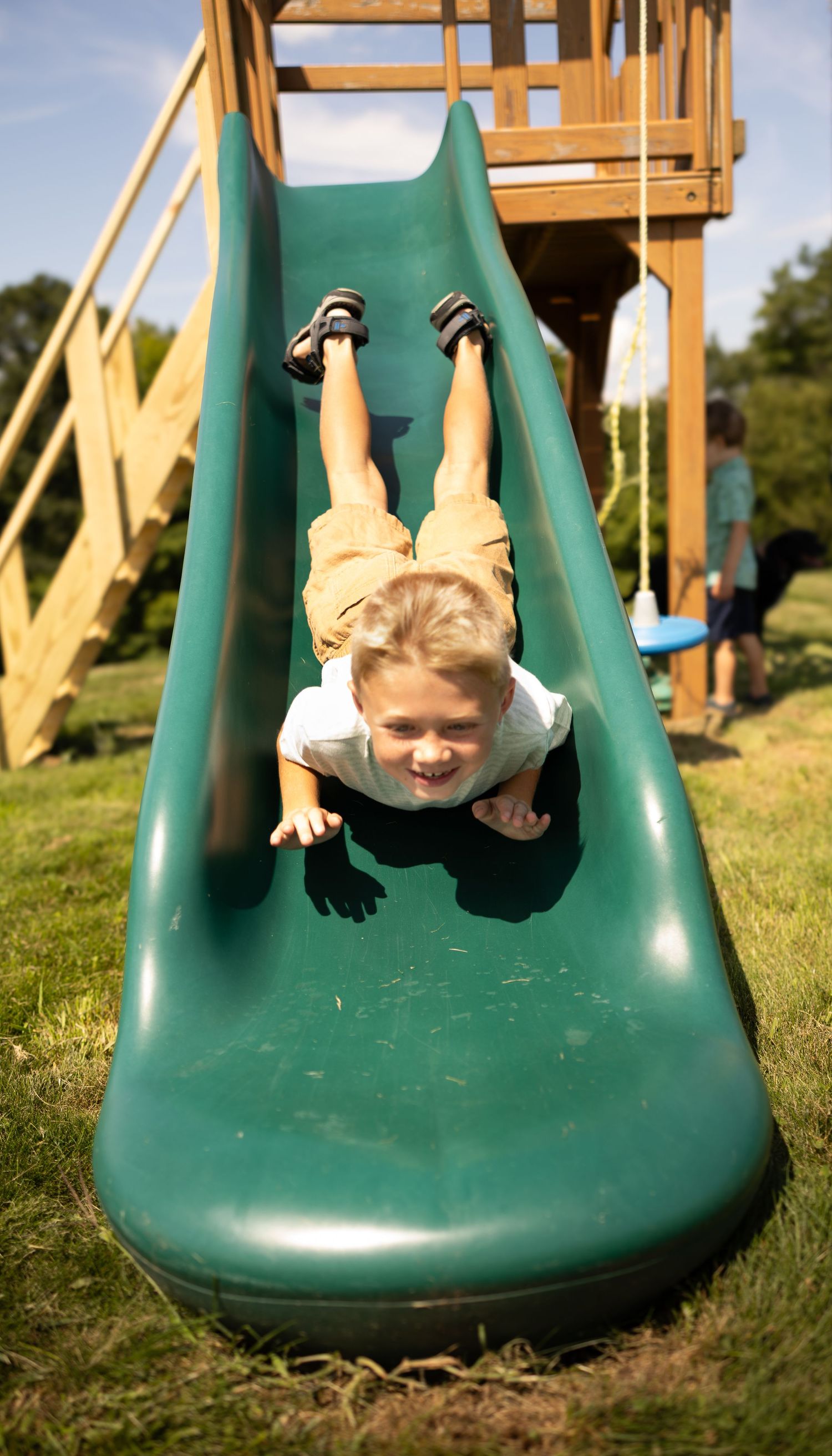 Young boy sliding headfirst down a green backyard playground slide on a sunny day.