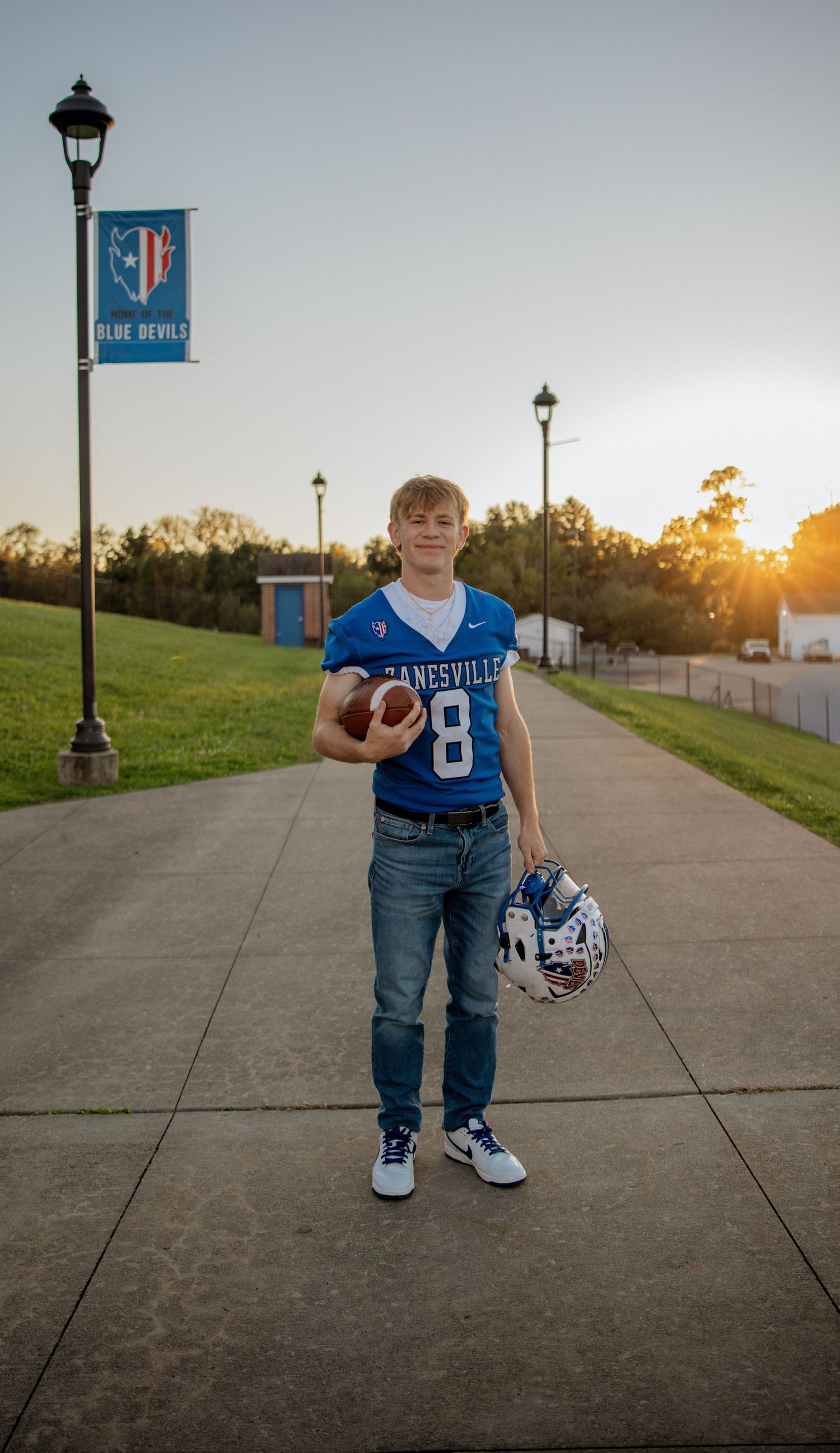 A young football player in a blue Zanesville Blue Devils jersey number 8 holds a ball and helmet at sunset.