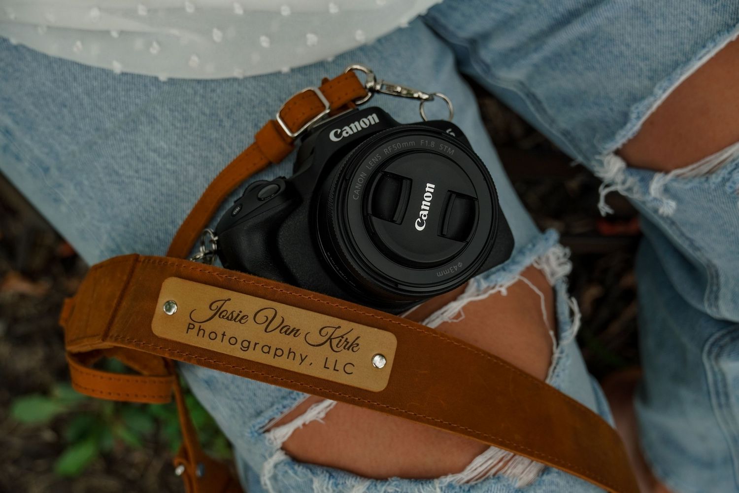 Canon camera with custom leather strap engraved Josie Van Kirk Photography LLC on denim lap.