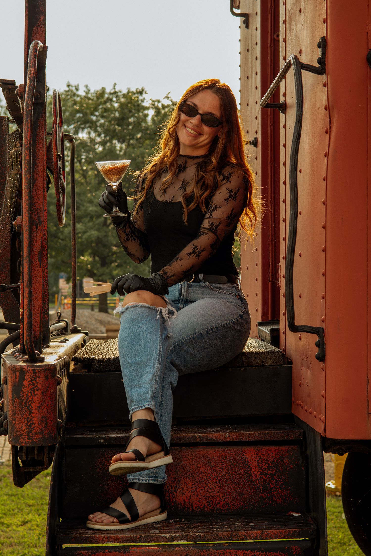 Smiling redhead woman in black lace top holding a cocktail glass while sitting on vintage train steps.