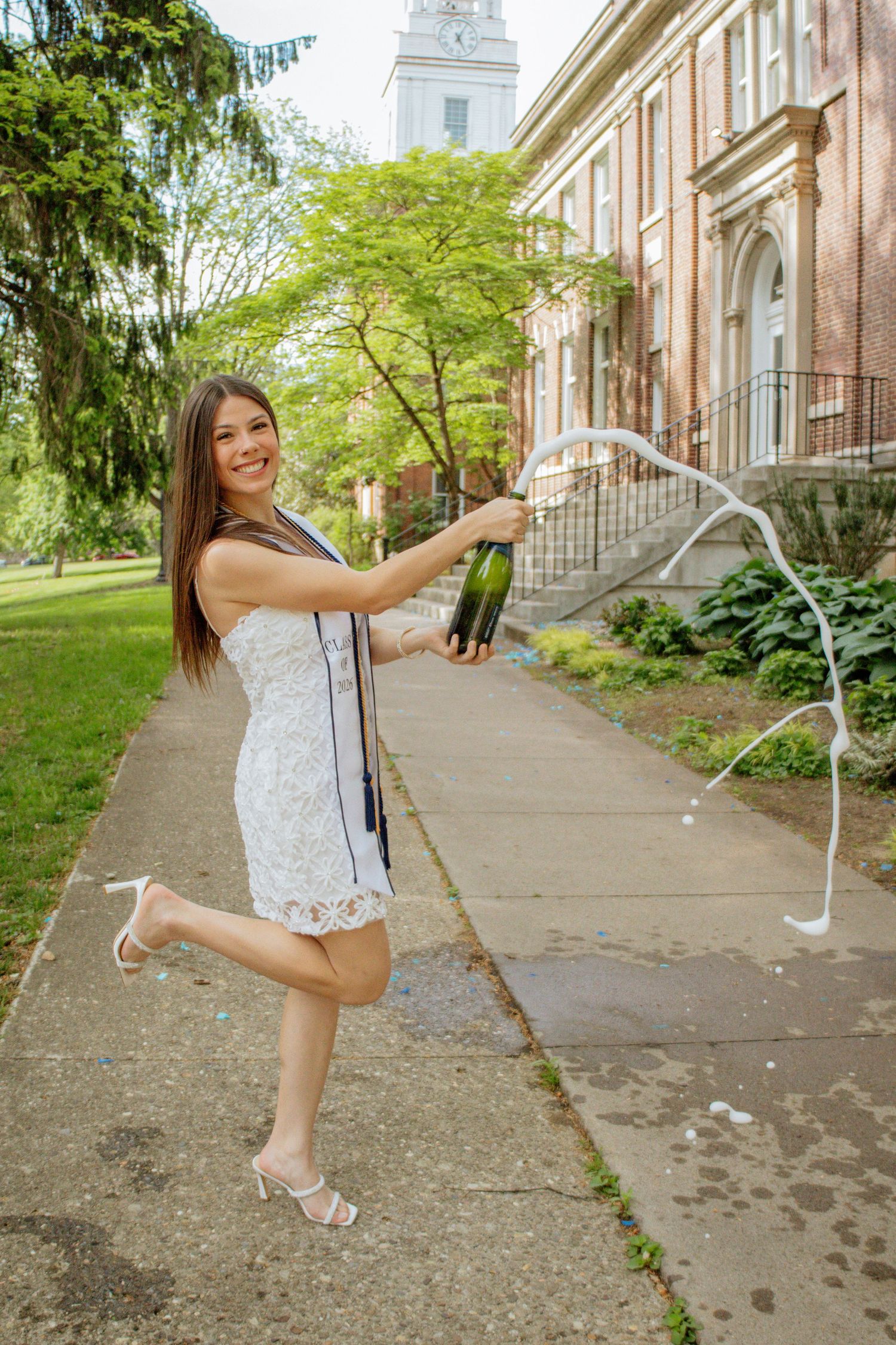 A young woman in a white dress pops champagne bottle on a college campus path during graduation.