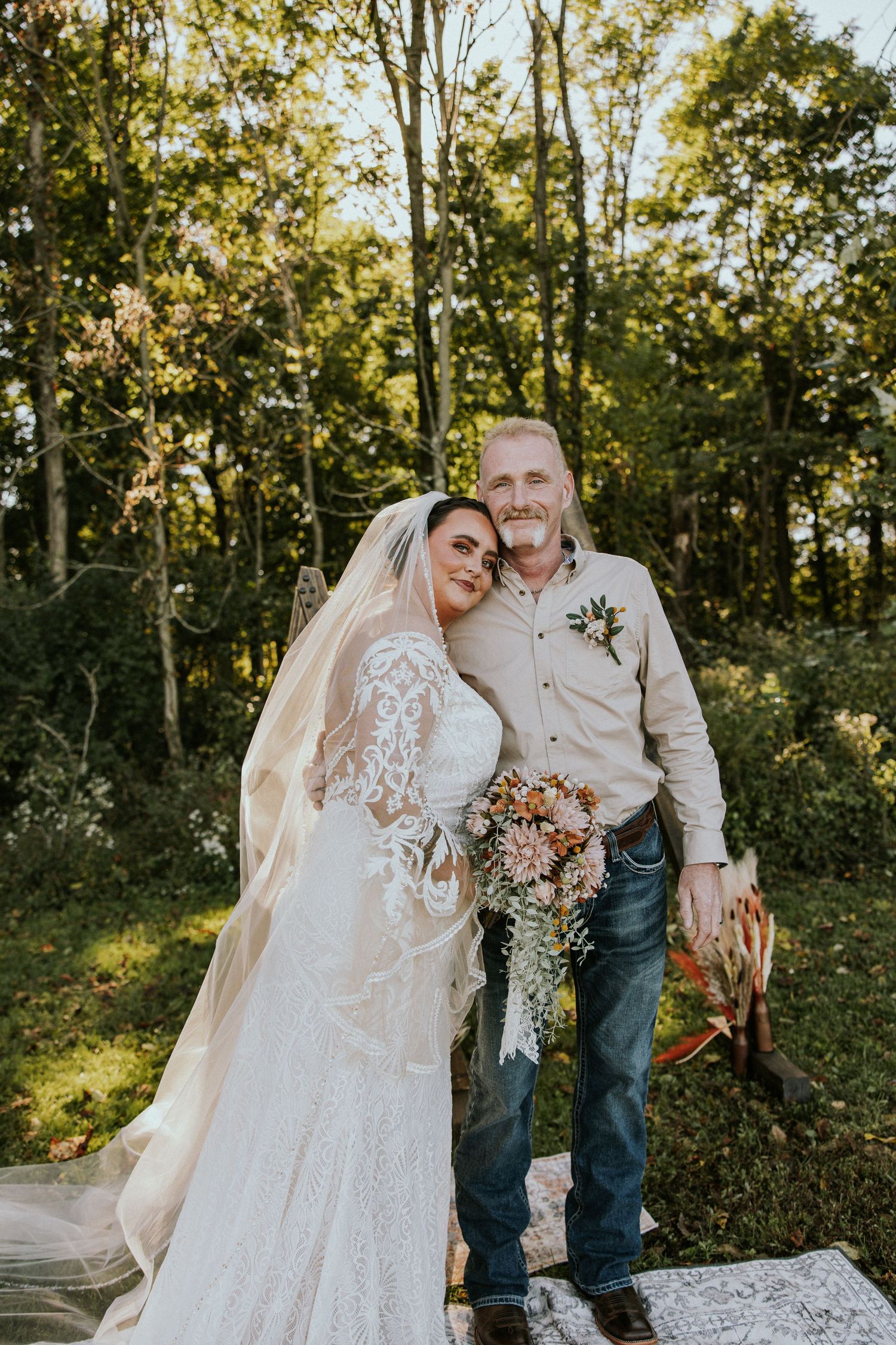 Bride in lace wedding gown and veil holds bouquet beside older man in beige shirt and jeans outdoors.