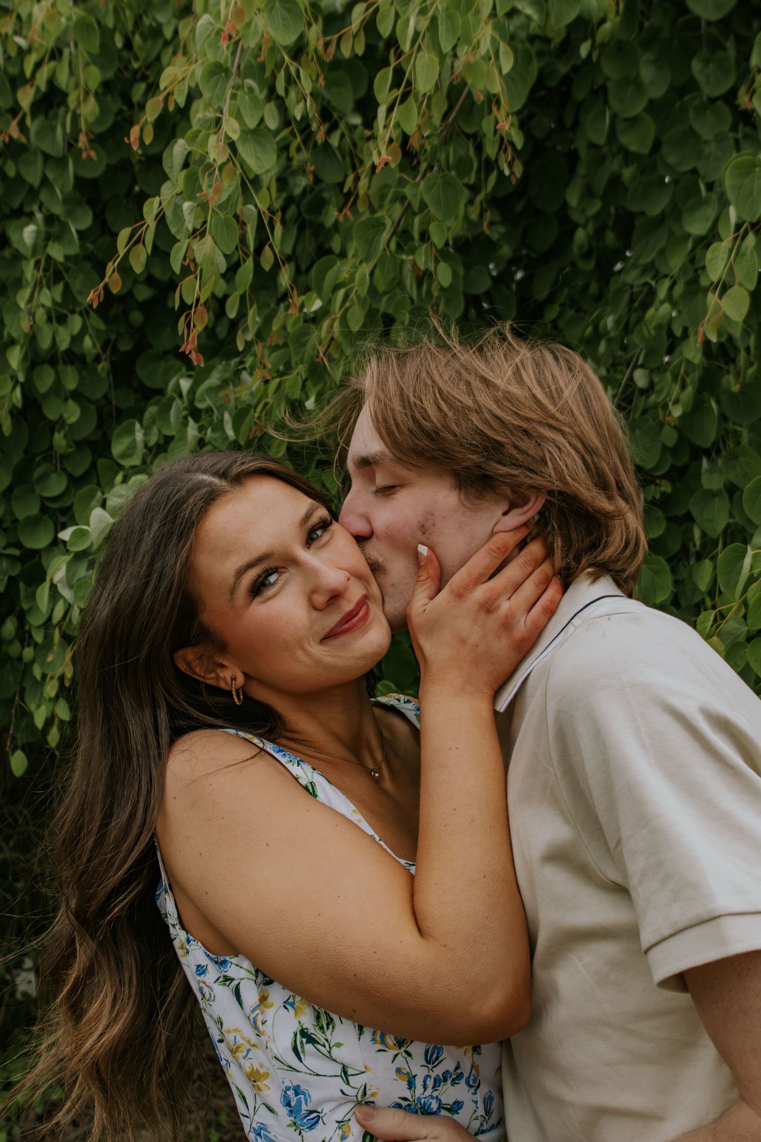 Couple sharing a tender kiss near a lush green ivy wall in a romantic outdoor portrait.