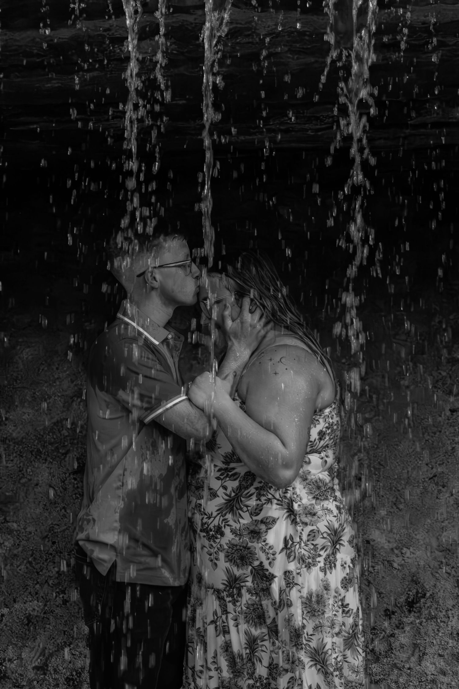 A couple shares a passionate kiss beneath cascading waterfall streams in black and white.