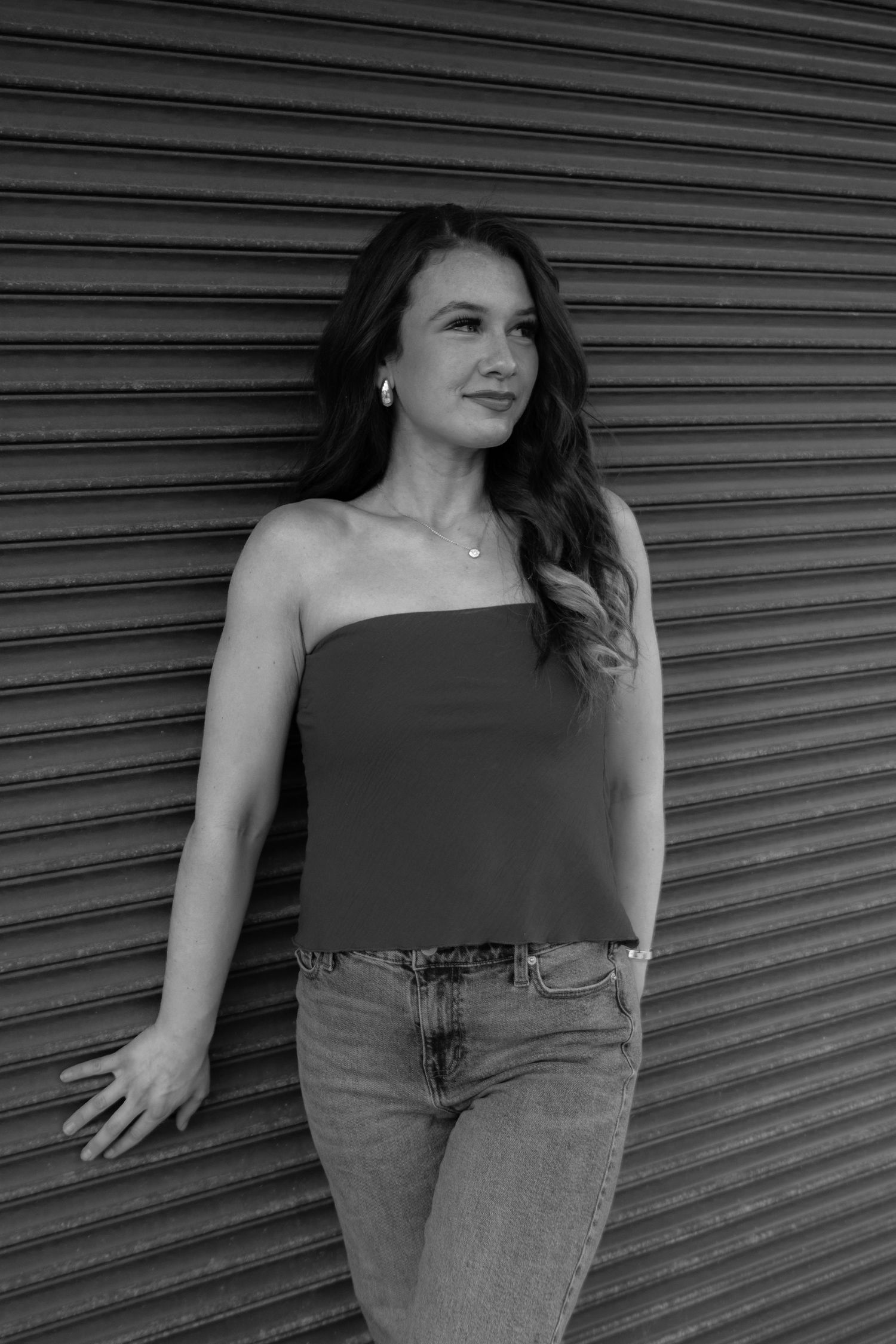 Black and white portrait of a brunette woman in a tube top and jeans leaning against a metal shutter wall.