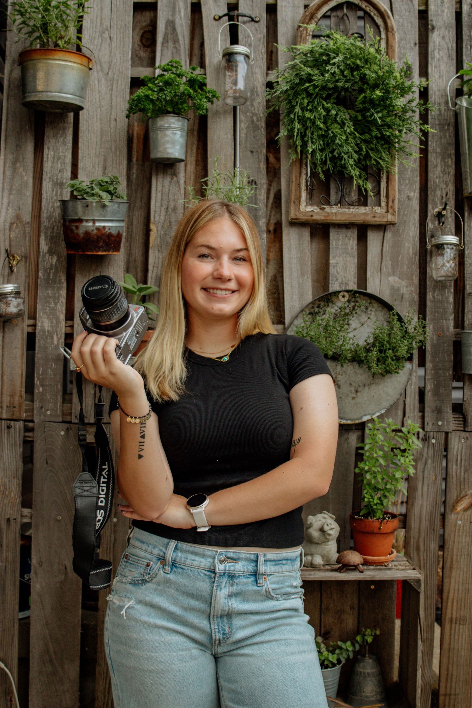 Smiling blonde photographer holding a camera outdoors in front of a rustic wooden wall with hanging potted plants.