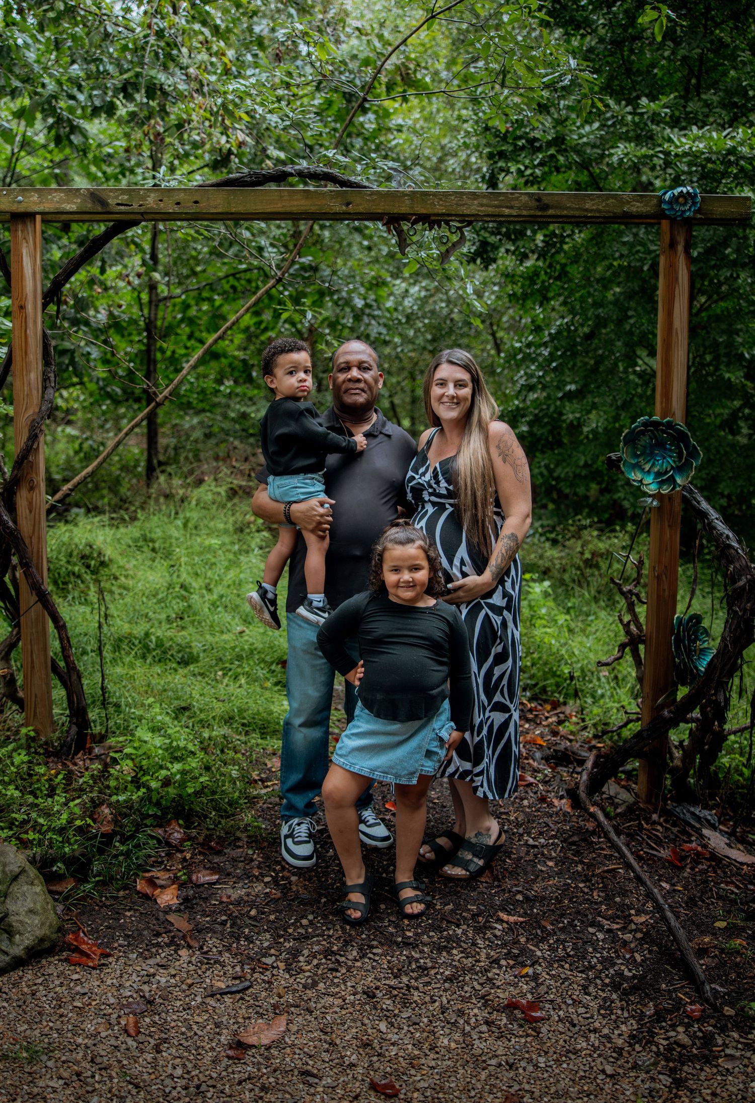 Family of four posing under rustic wooden arch surrounded by lush green trees in outdoor nature setting.