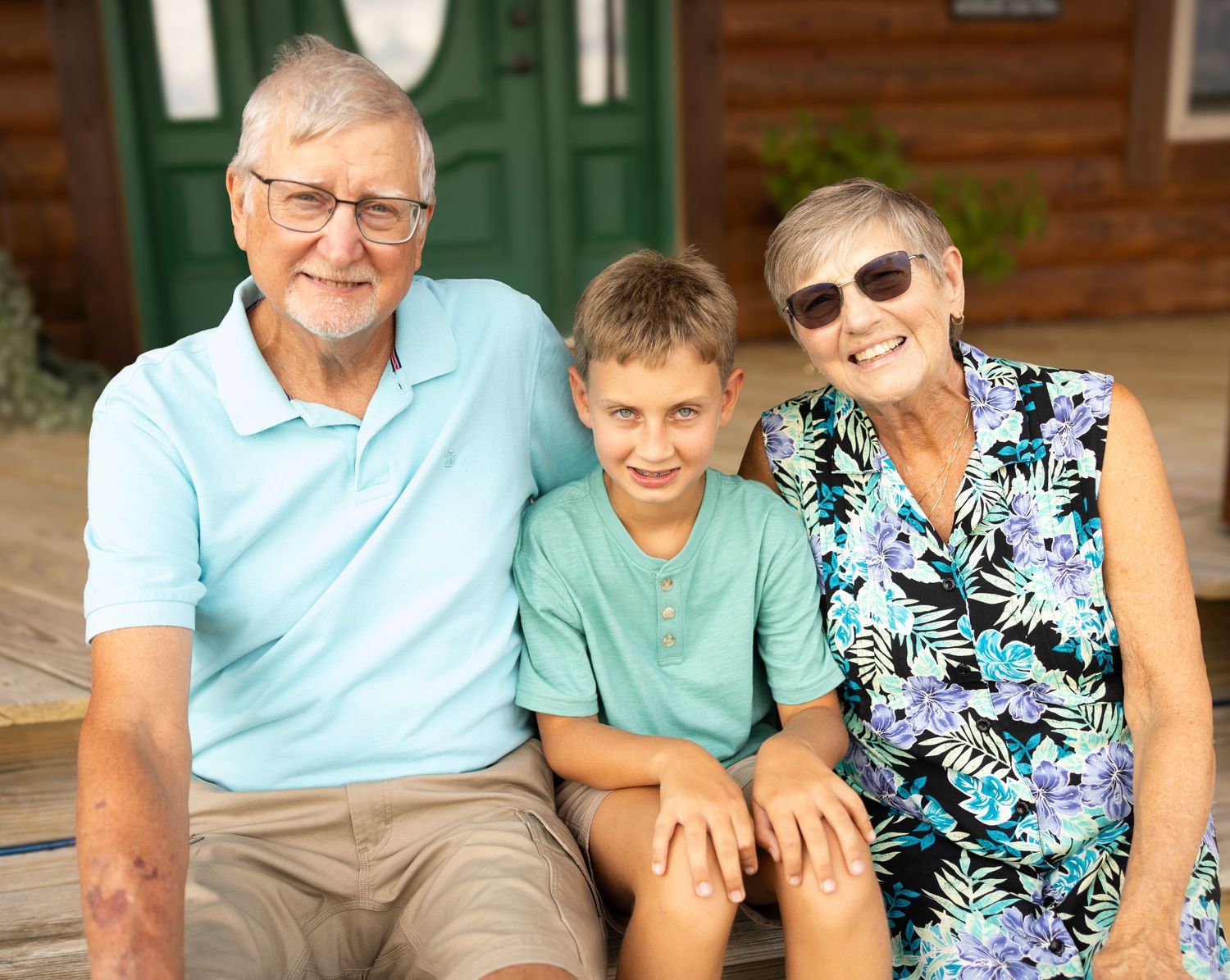 Grandparents and grandson smiling together on porch steps, senior couple with boy in matching teal shirts outdoors.