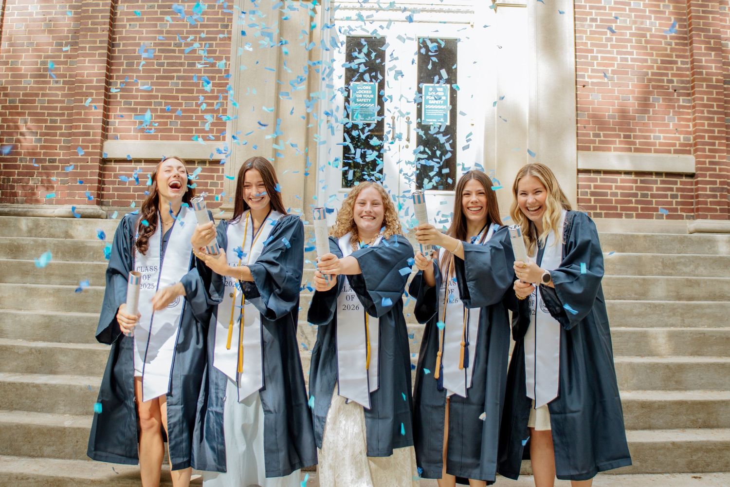 Five female graduates in navy caps and gowns celebrate with confetti outside a brick university building.