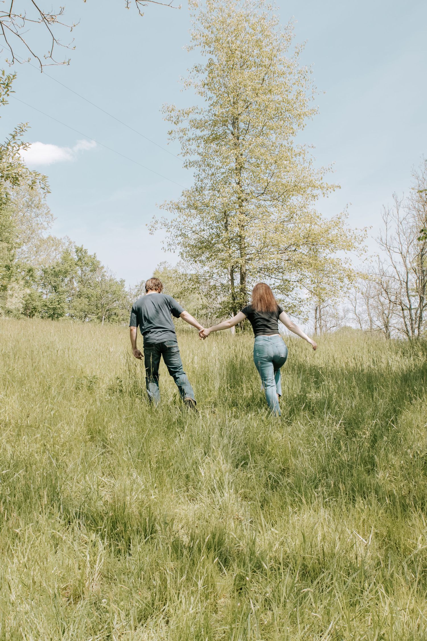 Couple holding hands walking through a lush green meadow with trees under a blue sky.
