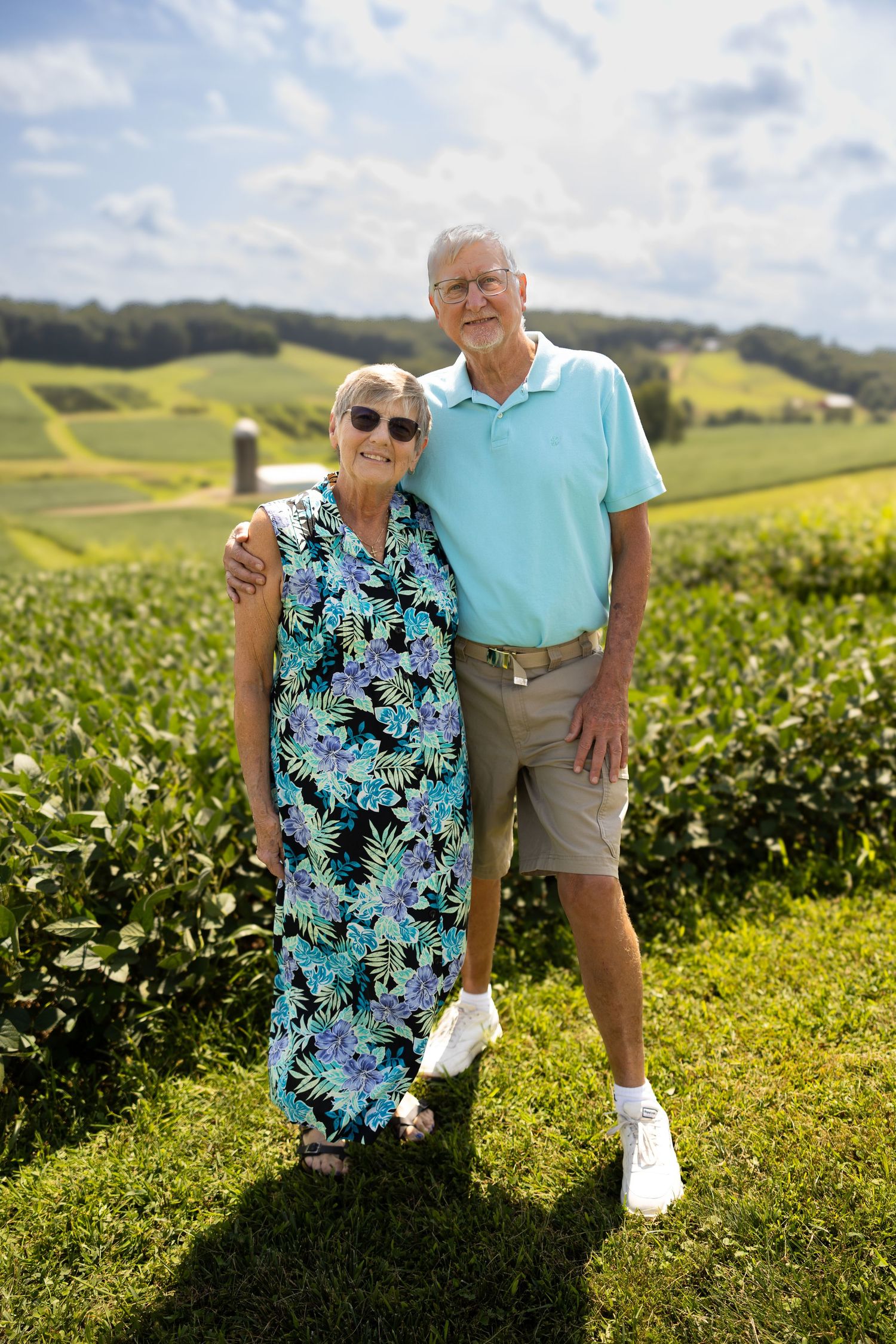 Elderly couple smiling together outdoors, standing in front of lush green farmland on a sunny day.