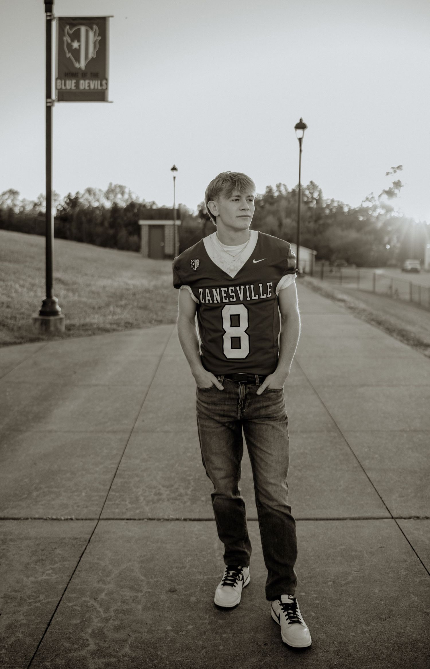 Black and white photo of a teen in a Zanesville #8 football jersey and jeans posing on a sidewalk near Blue Devils banner.