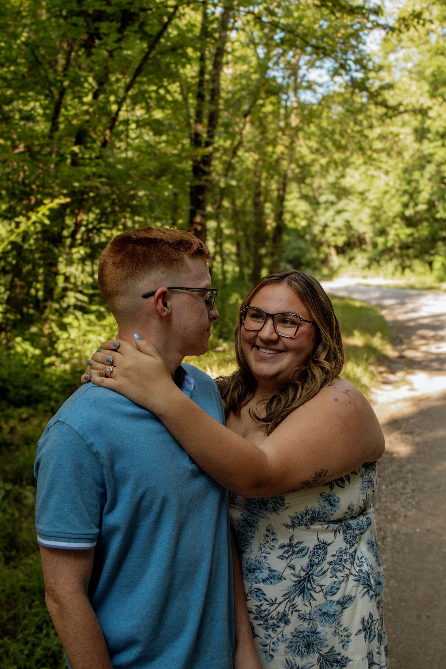 A smiling couple embraces on a sunlit forest path surrounded by lush green trees.