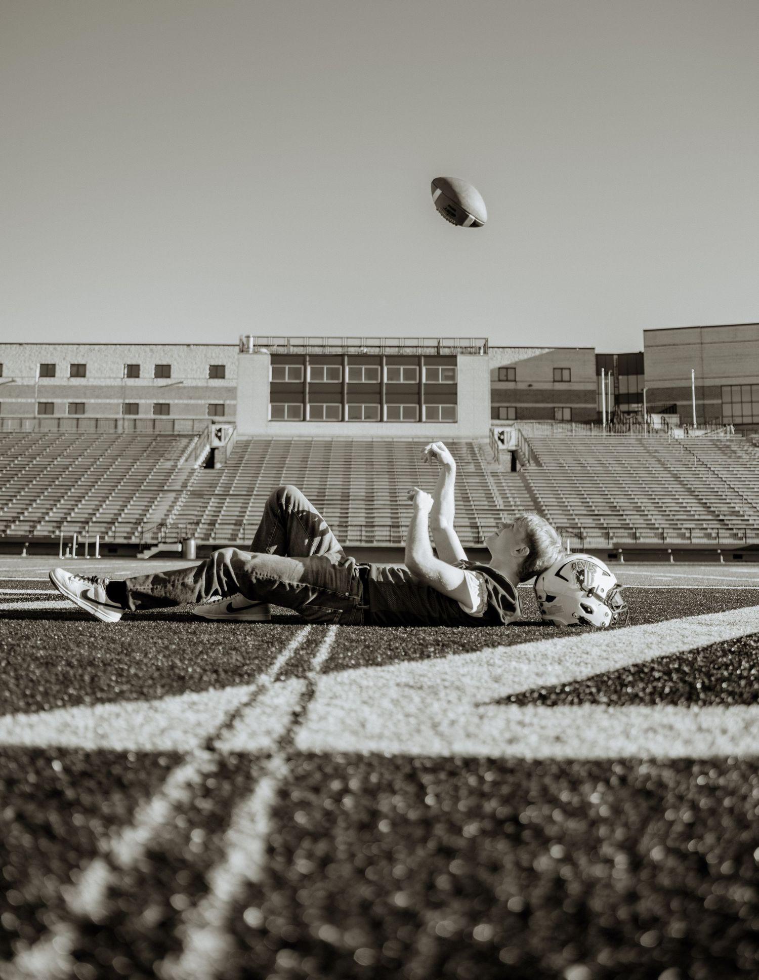 Person lying on football field tossing a ball in the air at an empty stadium, black and white photo.