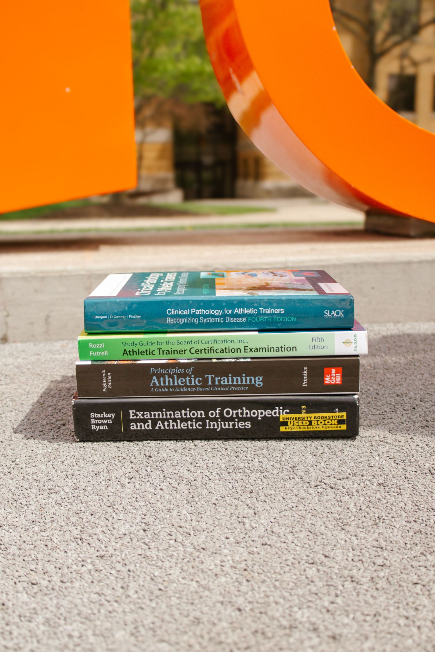Stack of athletic training textbooks on pavement near orange sculpture outdoors.