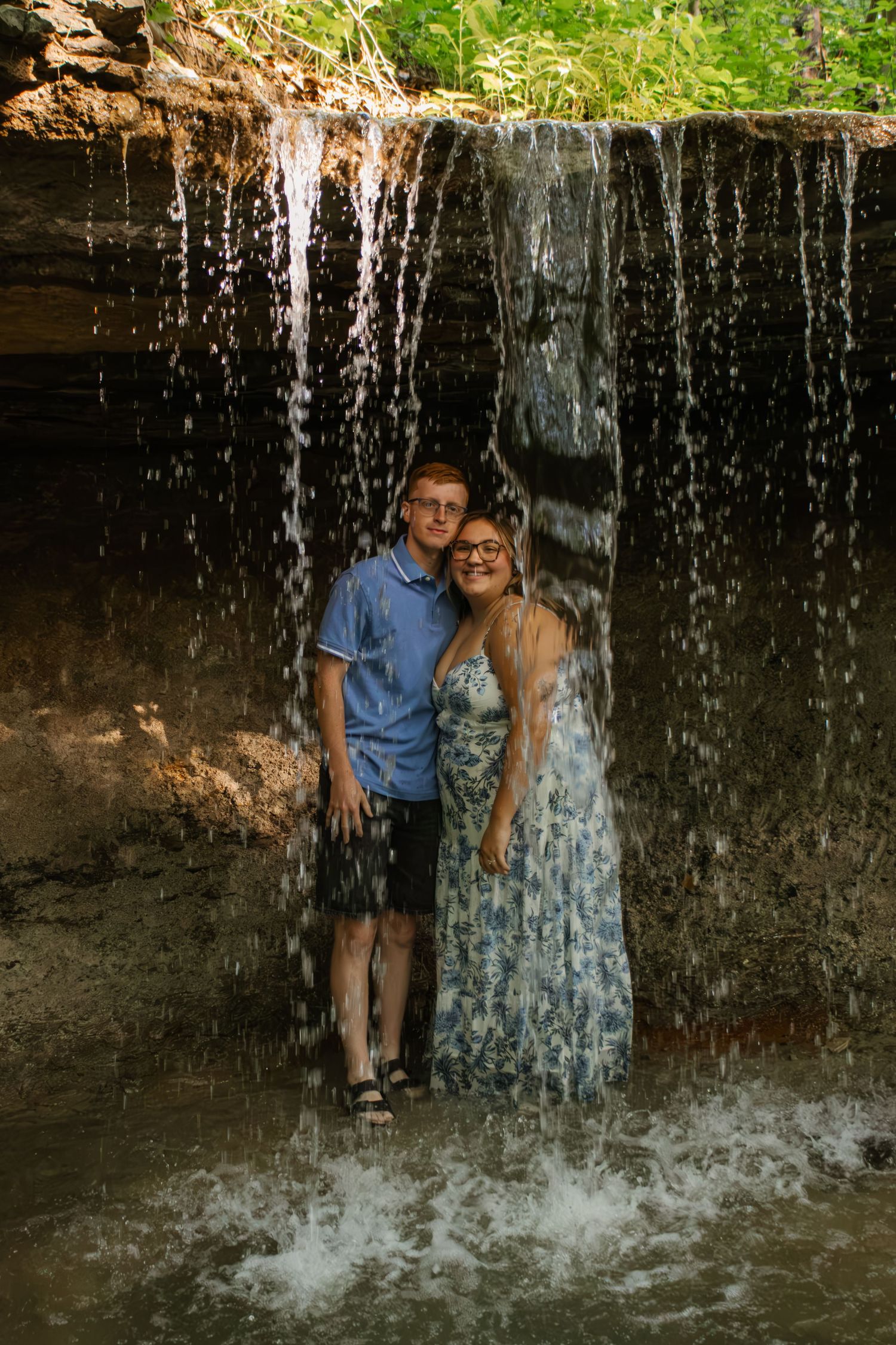 Happy couple embracing under a natural waterfall in a rocky outdoor setting surrounded by greenery.