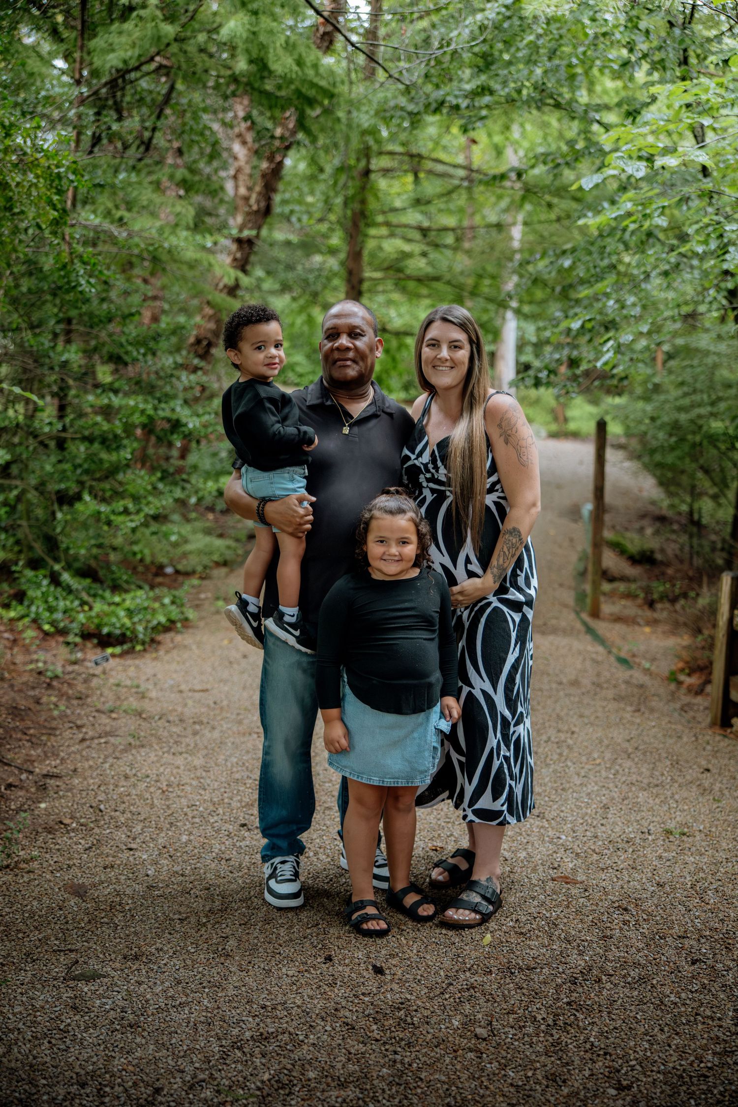 Family of four posing on a wooded trail, parents with two young children surrounded by lush green trees.
