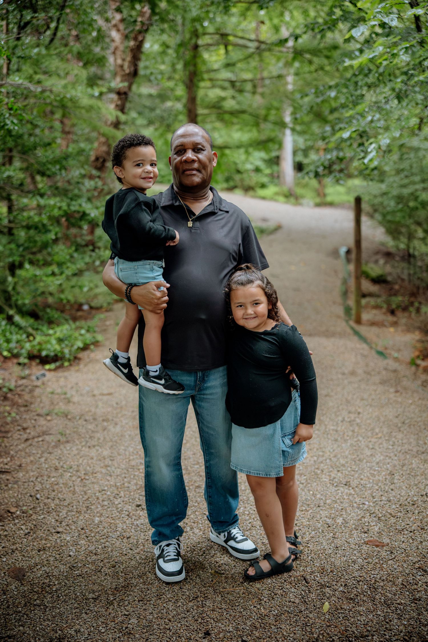 A man holds a toddler while a young girl stands beside him on a wooded park path.