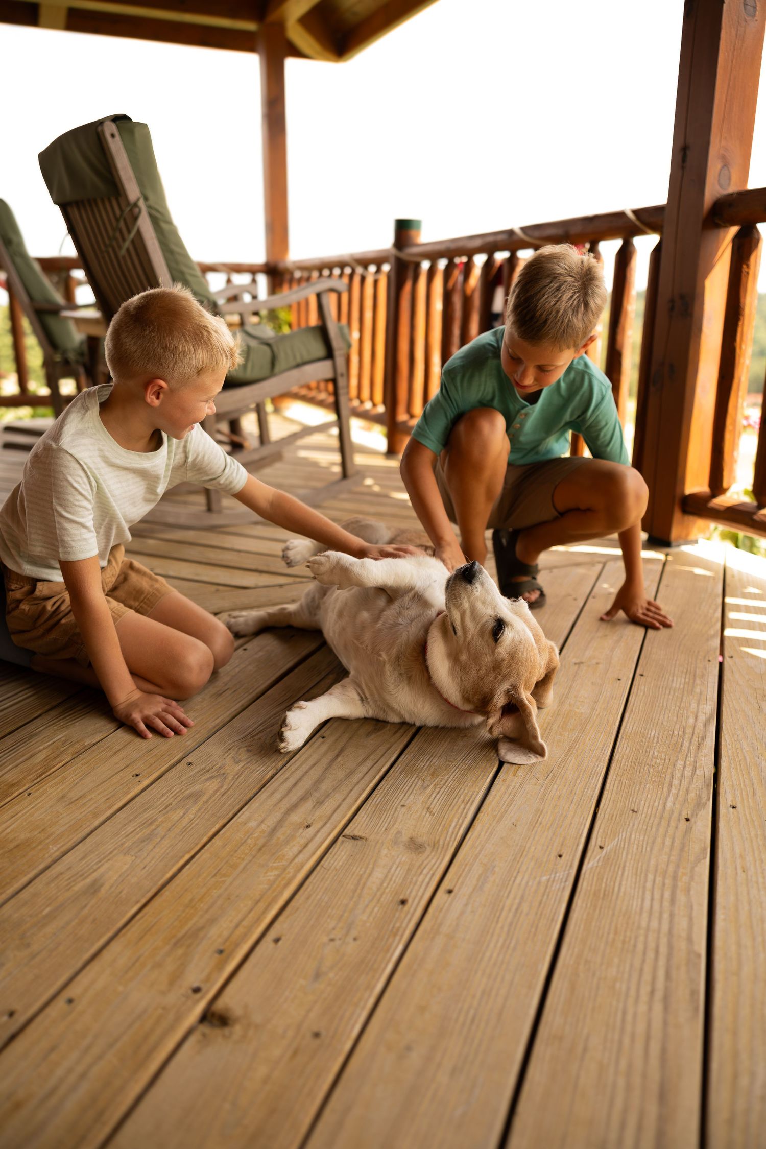 Two boys petting a golden dog on a wooden deck porch with rustic railings and outdoor chairs.