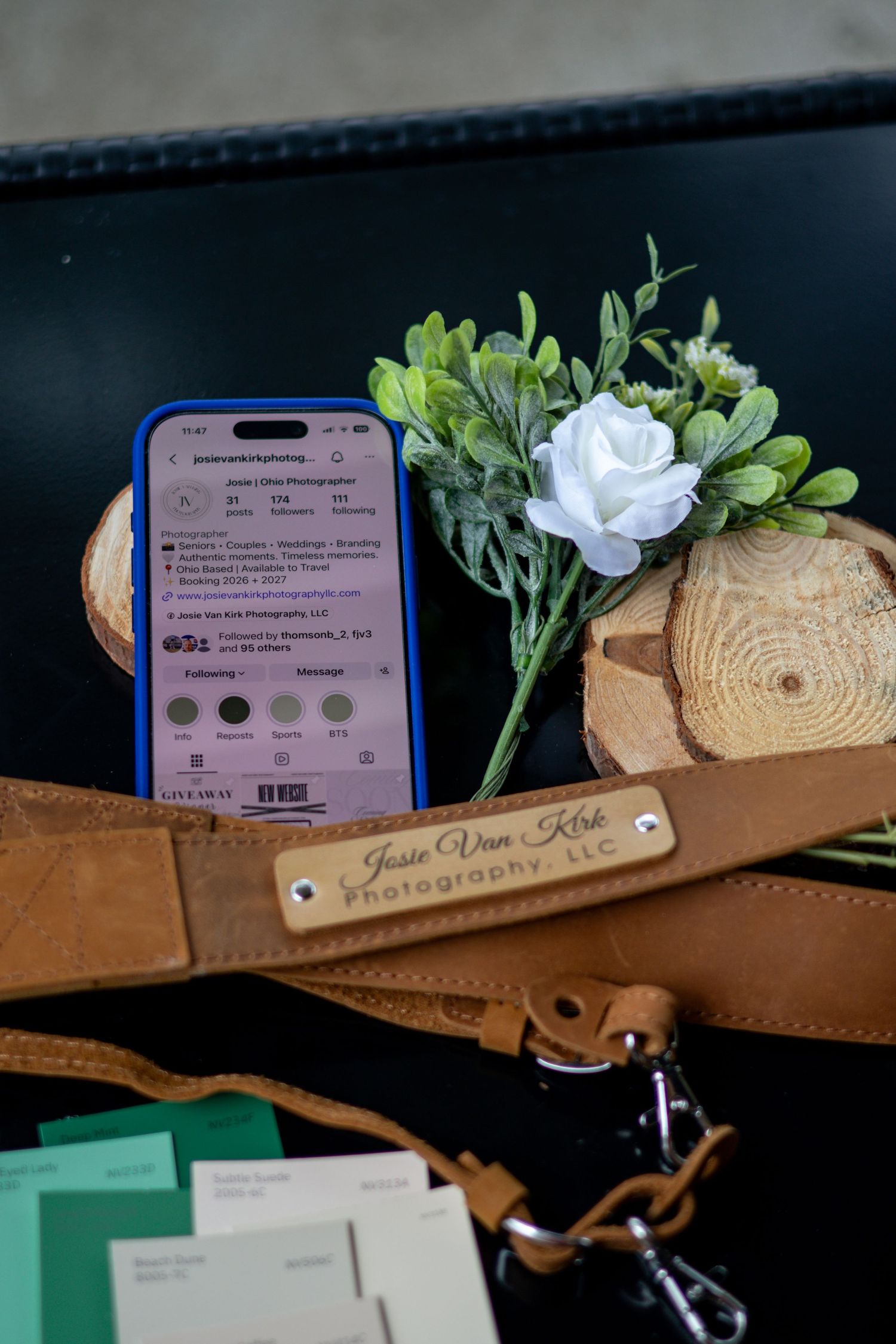 Jessie Van Kirk Photography LLC branded leather camera strap displayed with phone, flower, and wood slices.