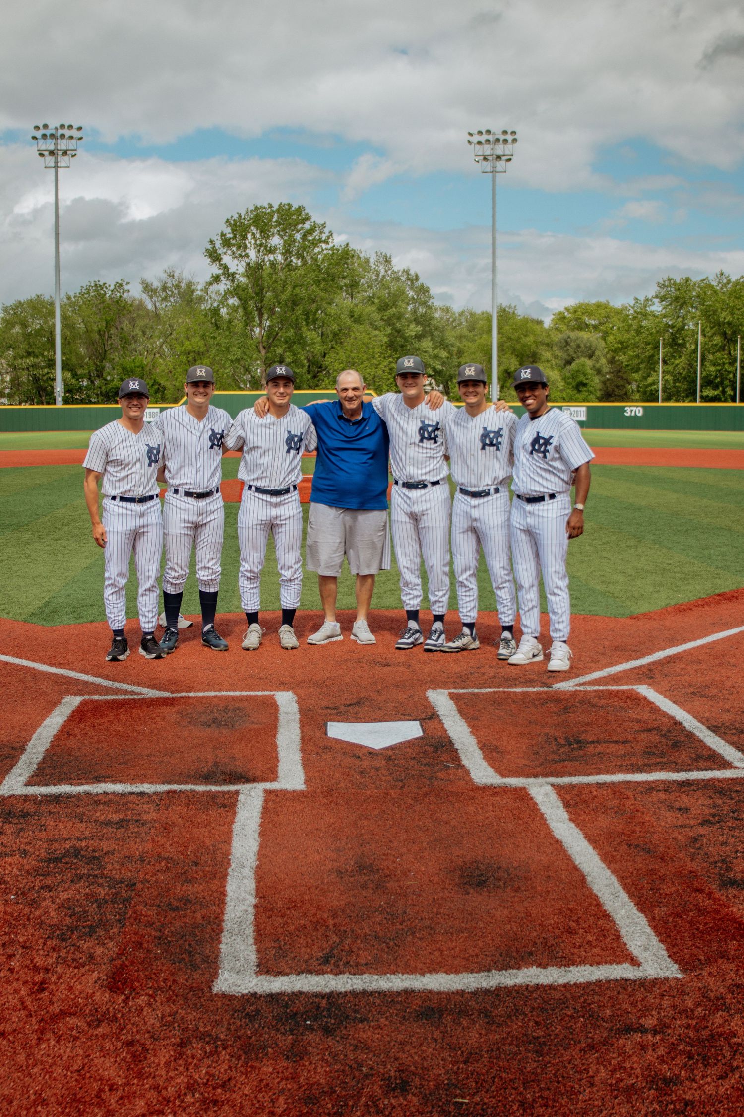 Baseball team seniors in uniforms posing with coach at home plate on red clay field.