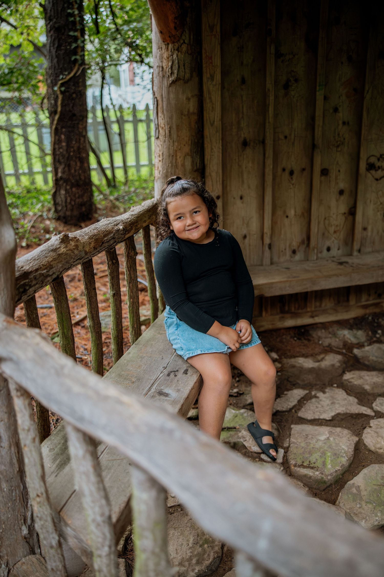Young girl in black top and denim skirt smiling on wooden porch steps of rustic cabin.