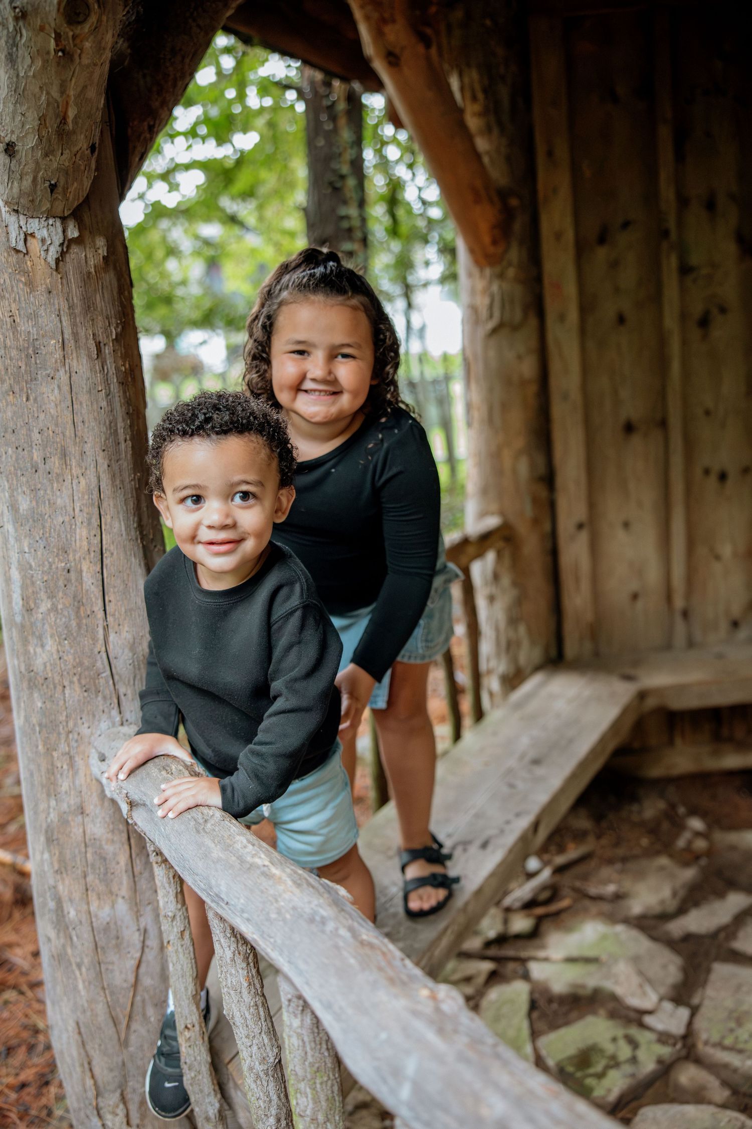 Two young children smiling in a rustic wooden treehouse, wearing matching black tops, enjoying outdoor playtime together.