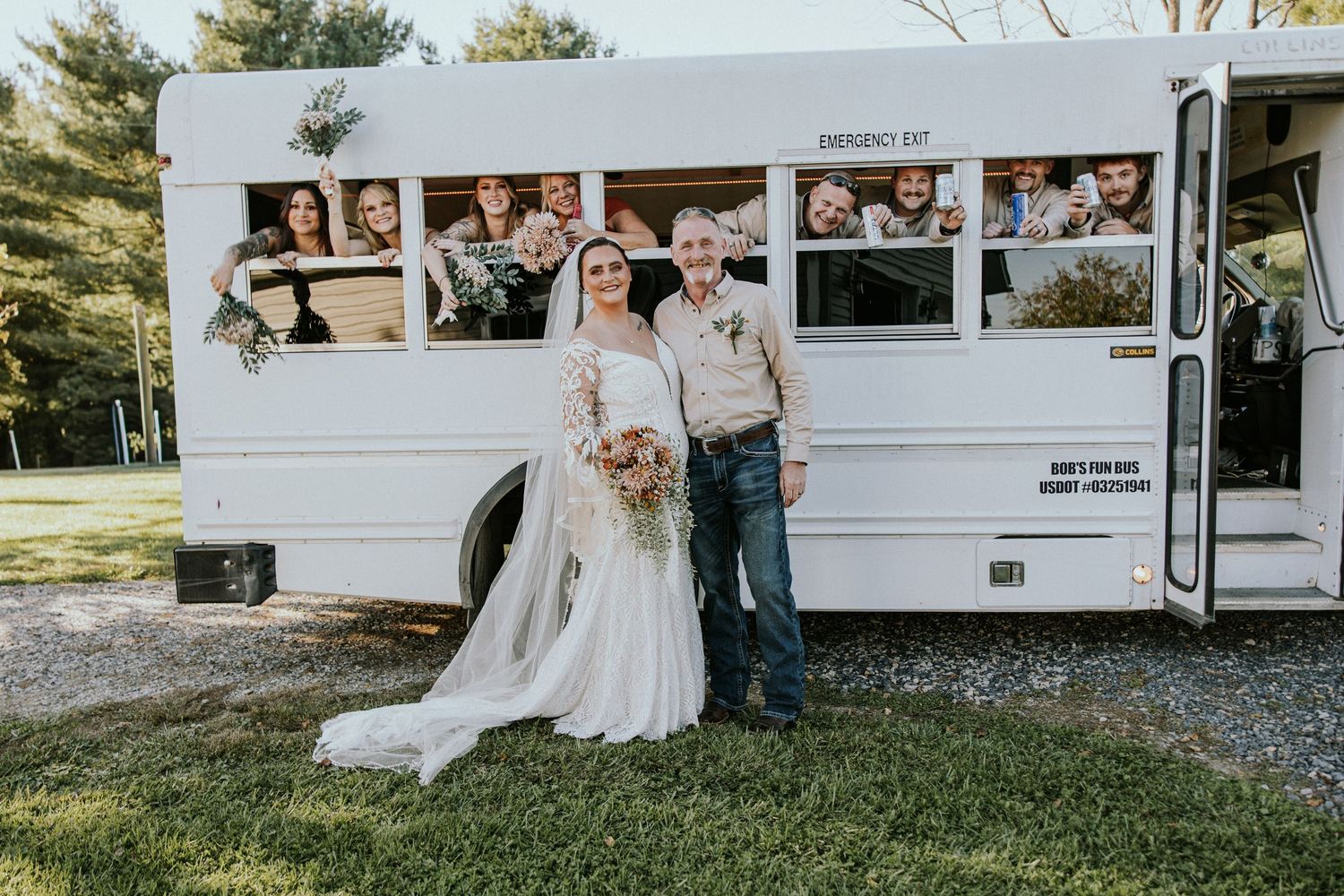 Bride and groom pose in front of a white vintage wedding bus with guests waving from windows on a sunny day.