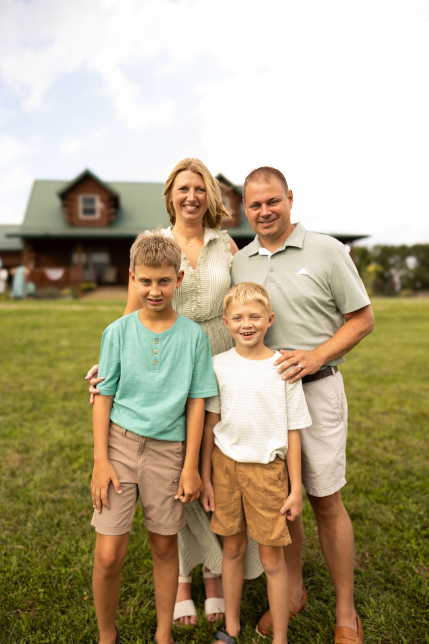 Happy family of four posing outdoors in front of a log cabin on a green lawn.
