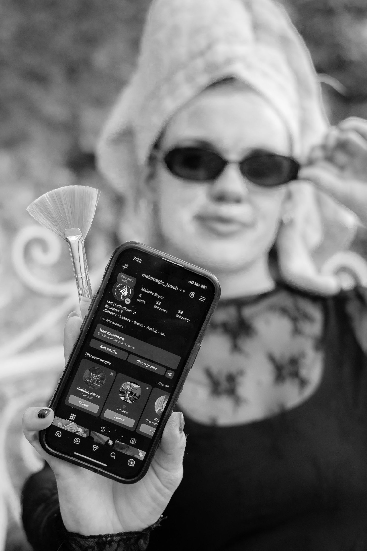 Black and white photo of blonde woman with towel turban holding phone showing Instagram profile page.