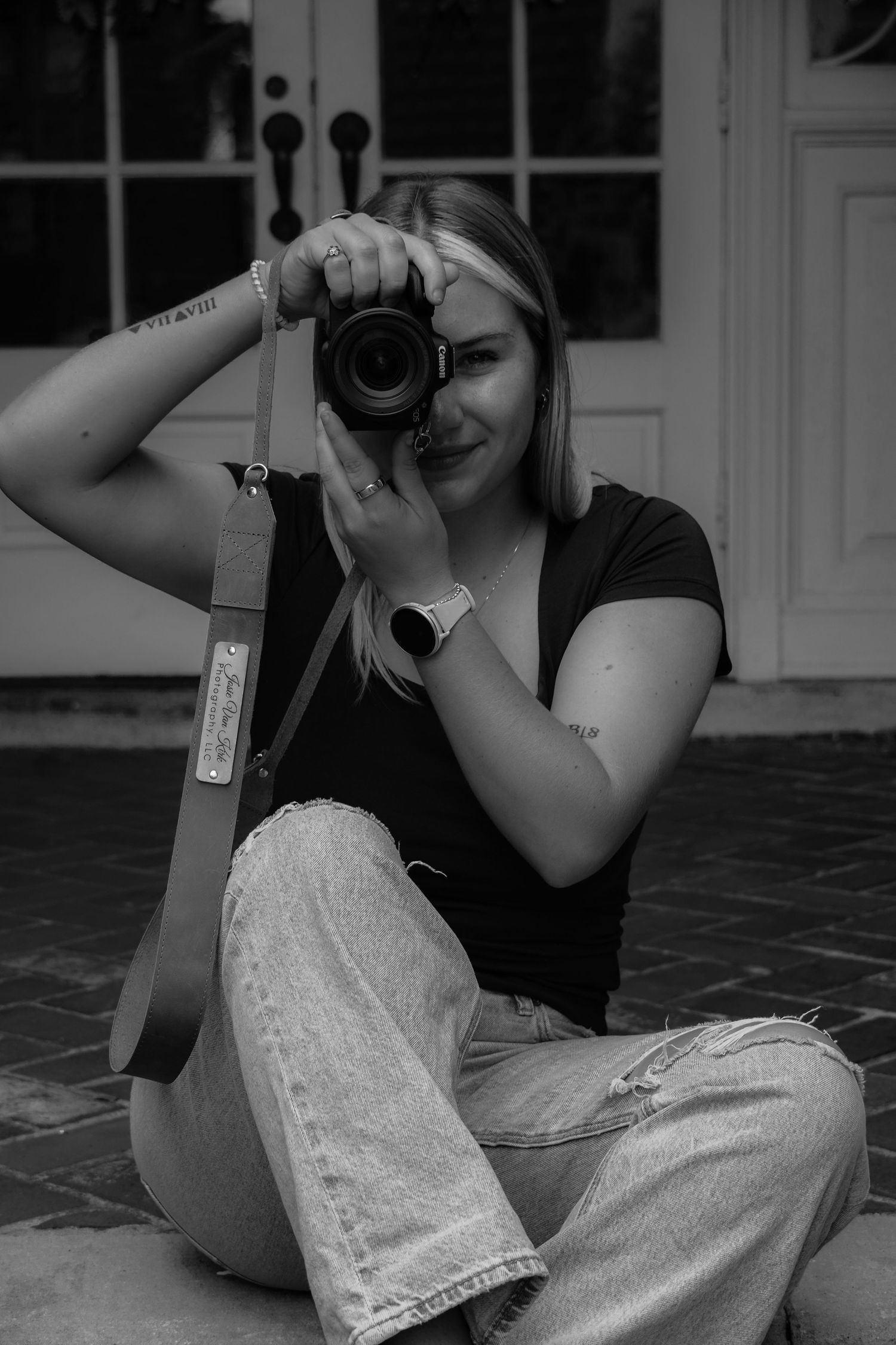 Black and white portrait of blonde female photographer sitting cross-legged, holding Canon camera up to her face.