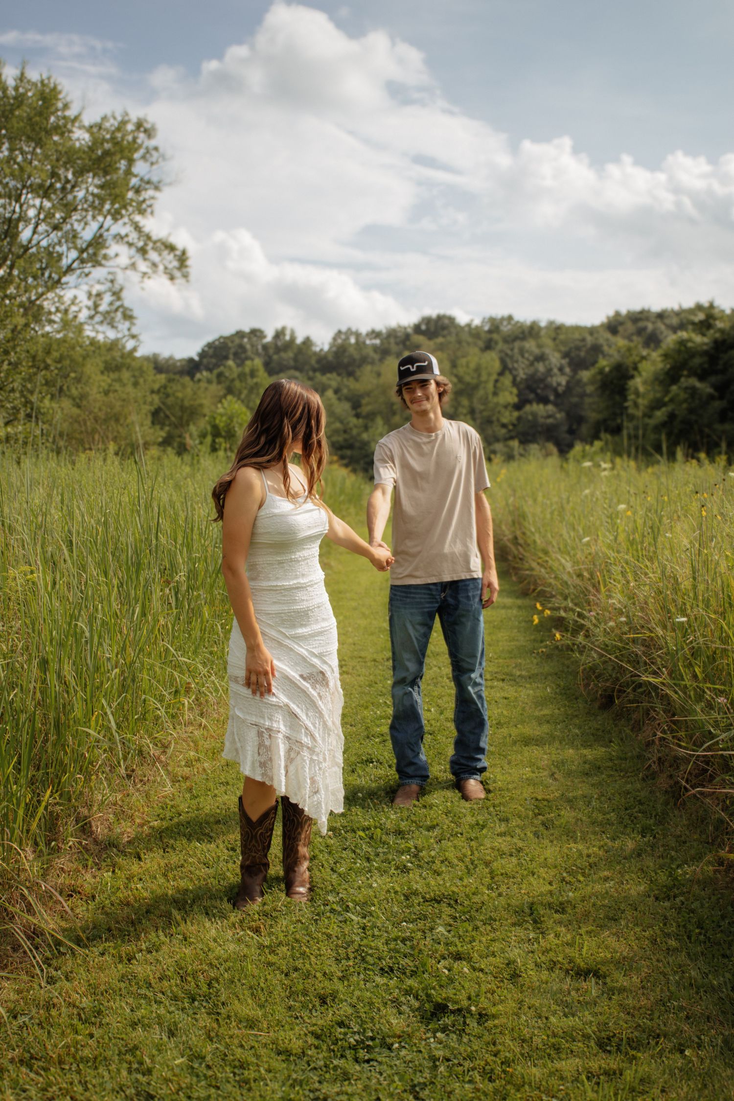 A young couple holds hands on a grassy path surrounded by lush green fields under a partly cloudy sky.