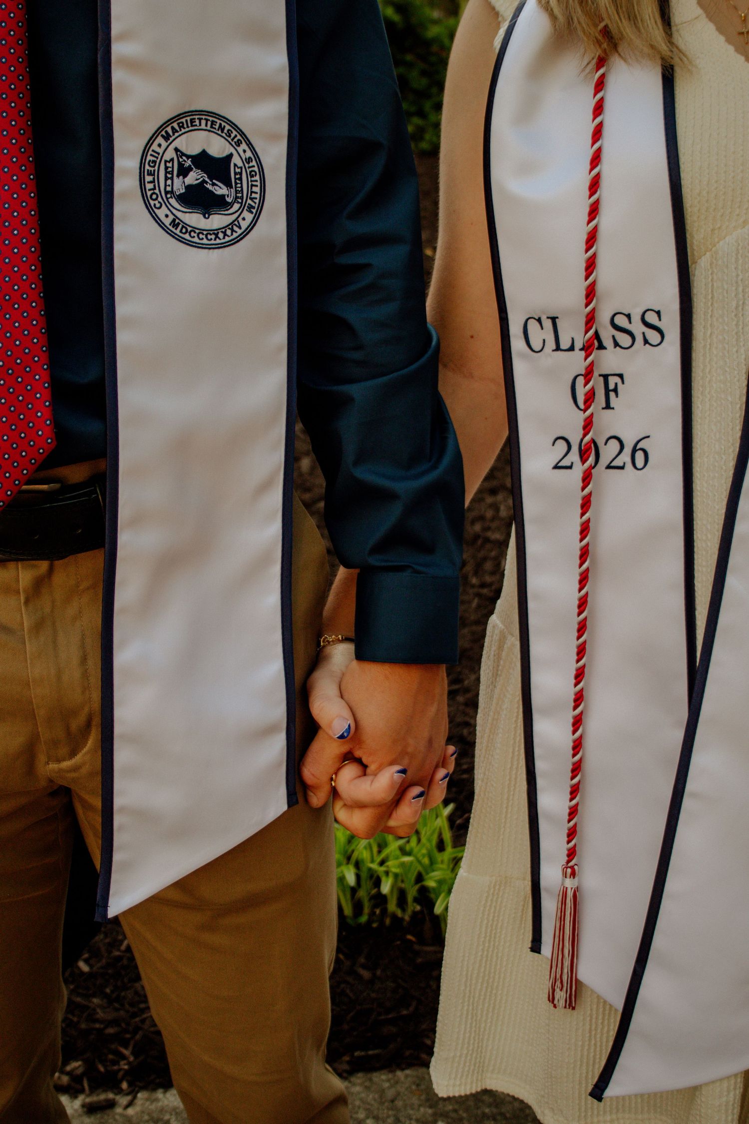 Close-up of two graduates holding hands wearing Class of 2026 graduation stoles on campus.