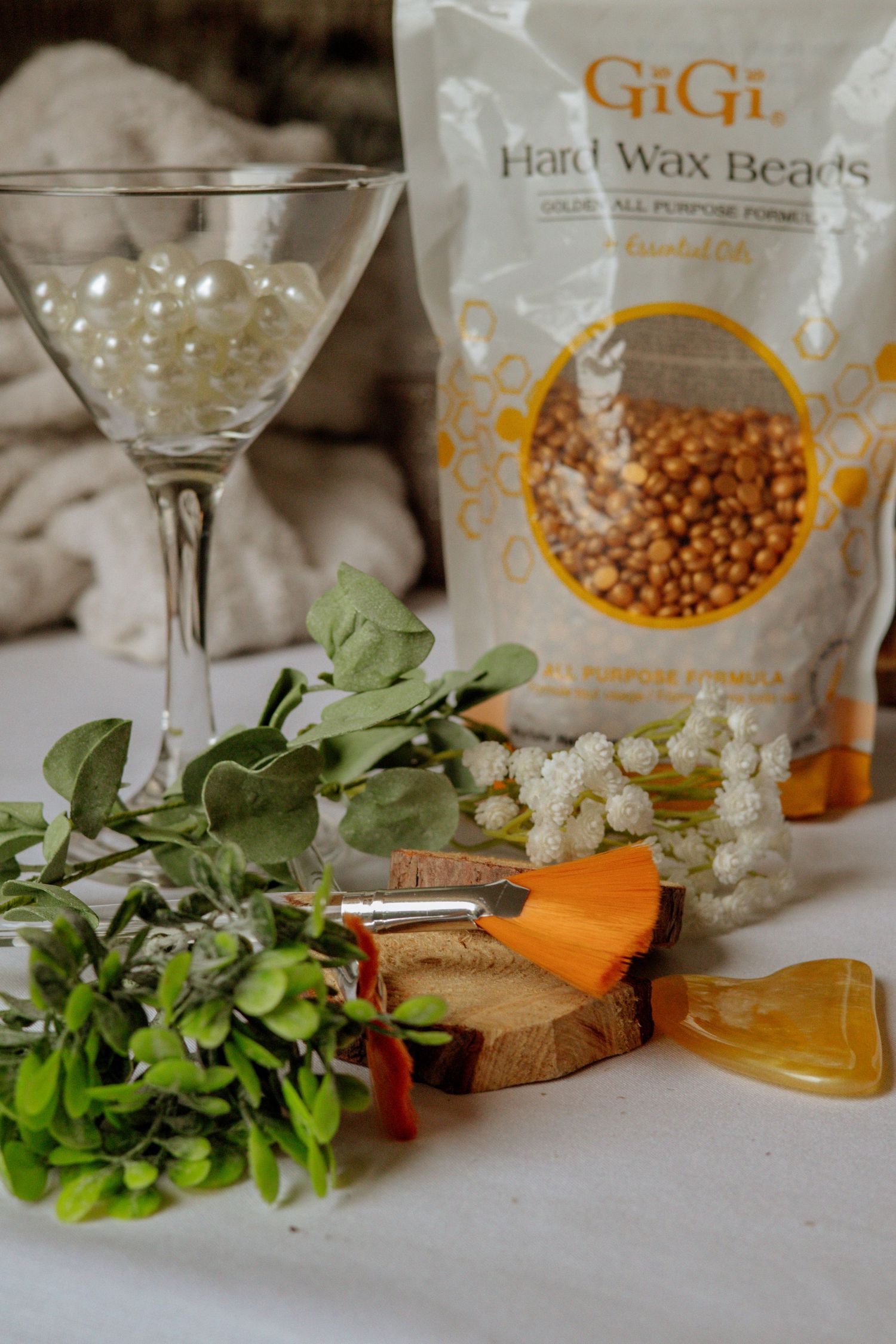 GiGi Hard Wax Beads bag displayed with martini glass, pearls, flowers, and beauty tools on white surface.