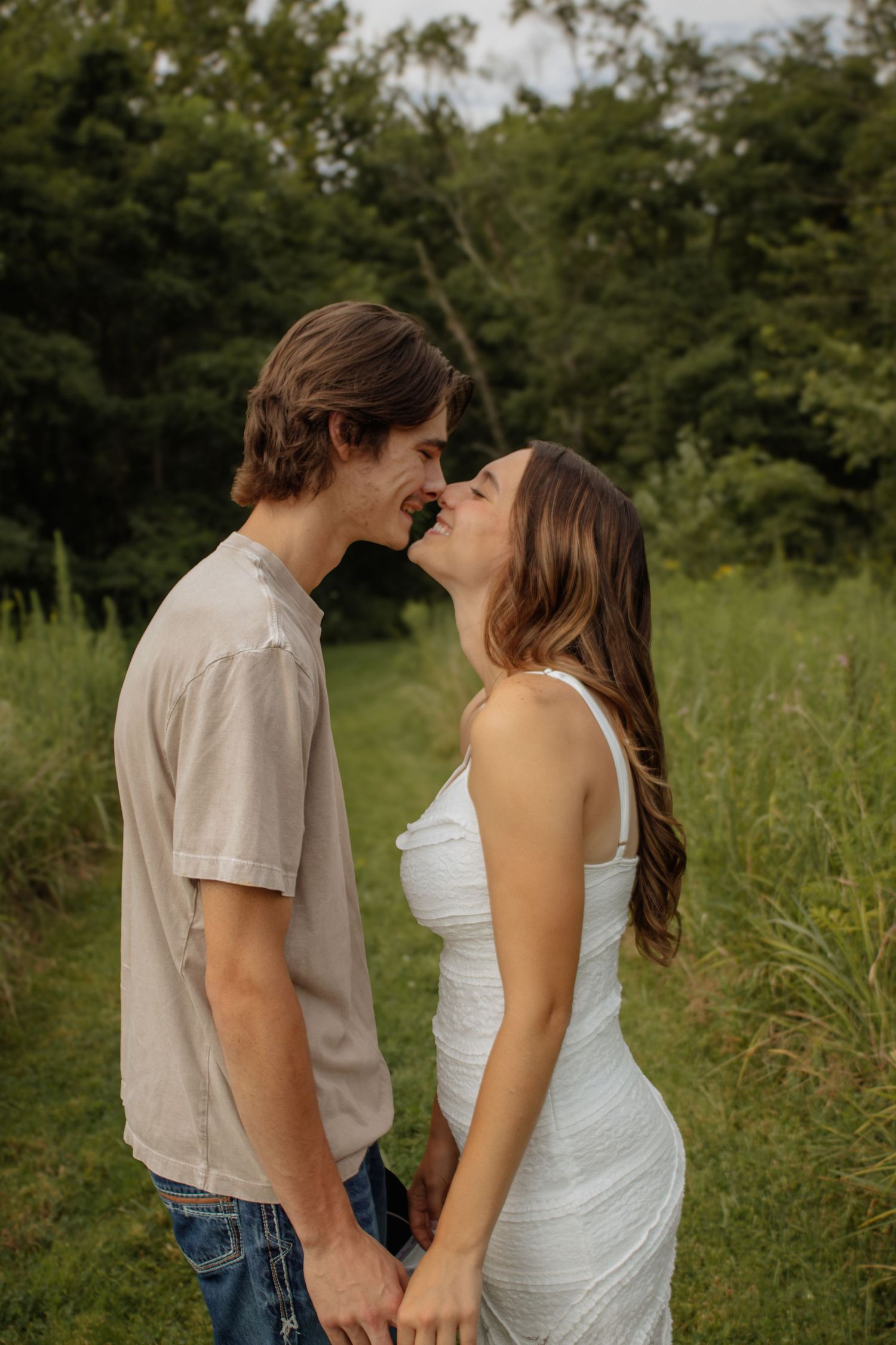 A couple shares a romantic kiss in a lush green field, woman in white dress holding hands with man in beige shirt.
