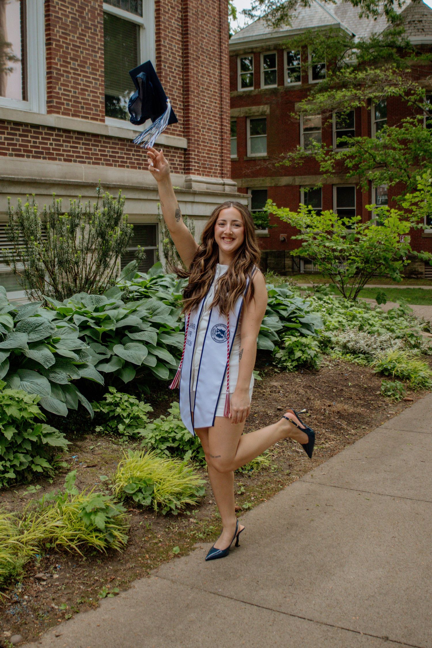 Happy graduate in white stole tossing cap outside brick college building.