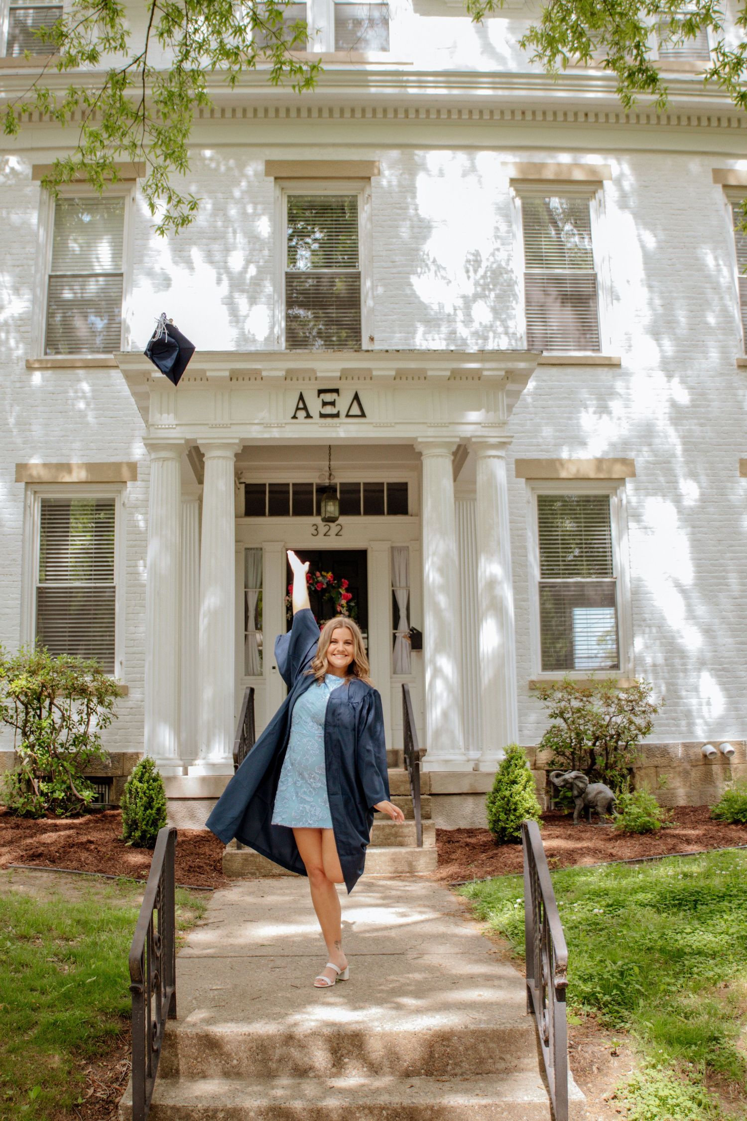 Joyful female graduate tossing cap in air outside Alpha Xi Delta sorority house in blue graduation gown.