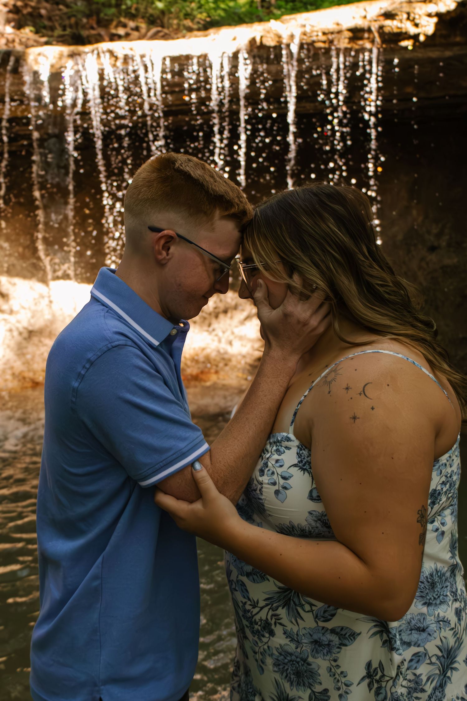 A romantic couple touches foreheads in front of a glittering waterfall in golden light.
