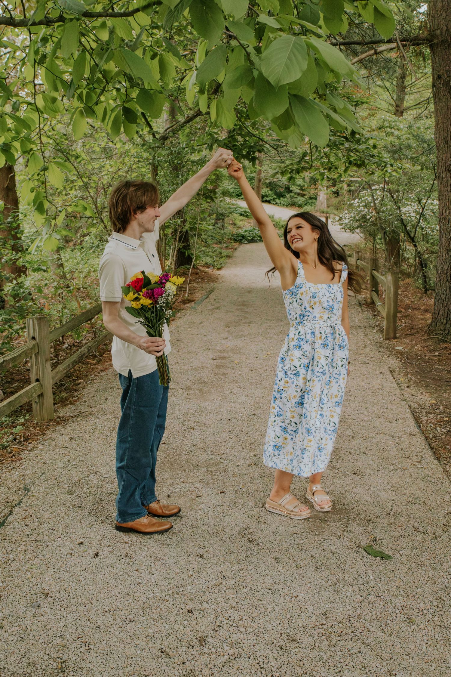 Joyful couple dancing on a gravel path surrounded by green trees, man holding bright flowers.