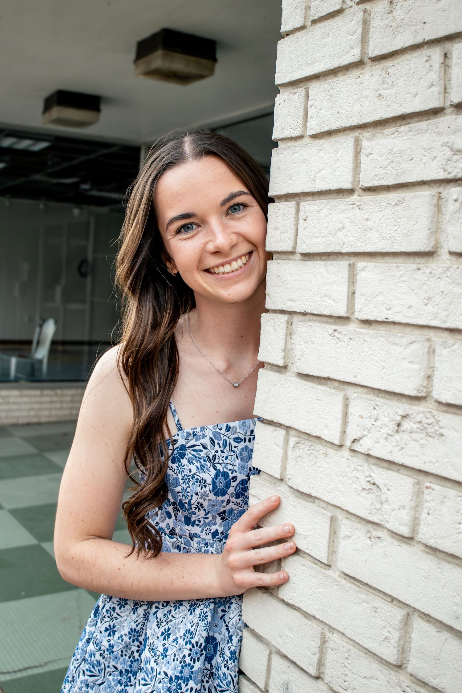 Young woman in floral blue dress smiling against white brick wall outdoors.