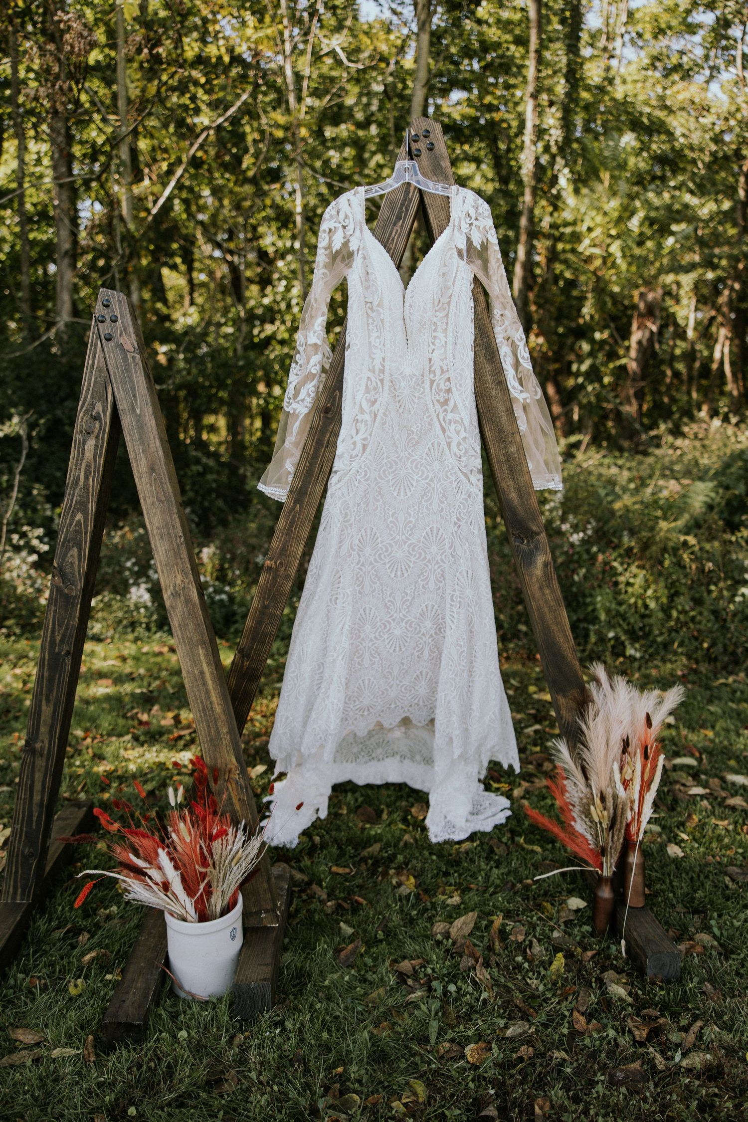 Boho lace wedding dress with long sleeves hanging on wooden arch outdoors, flanked by wildflower bouquets.