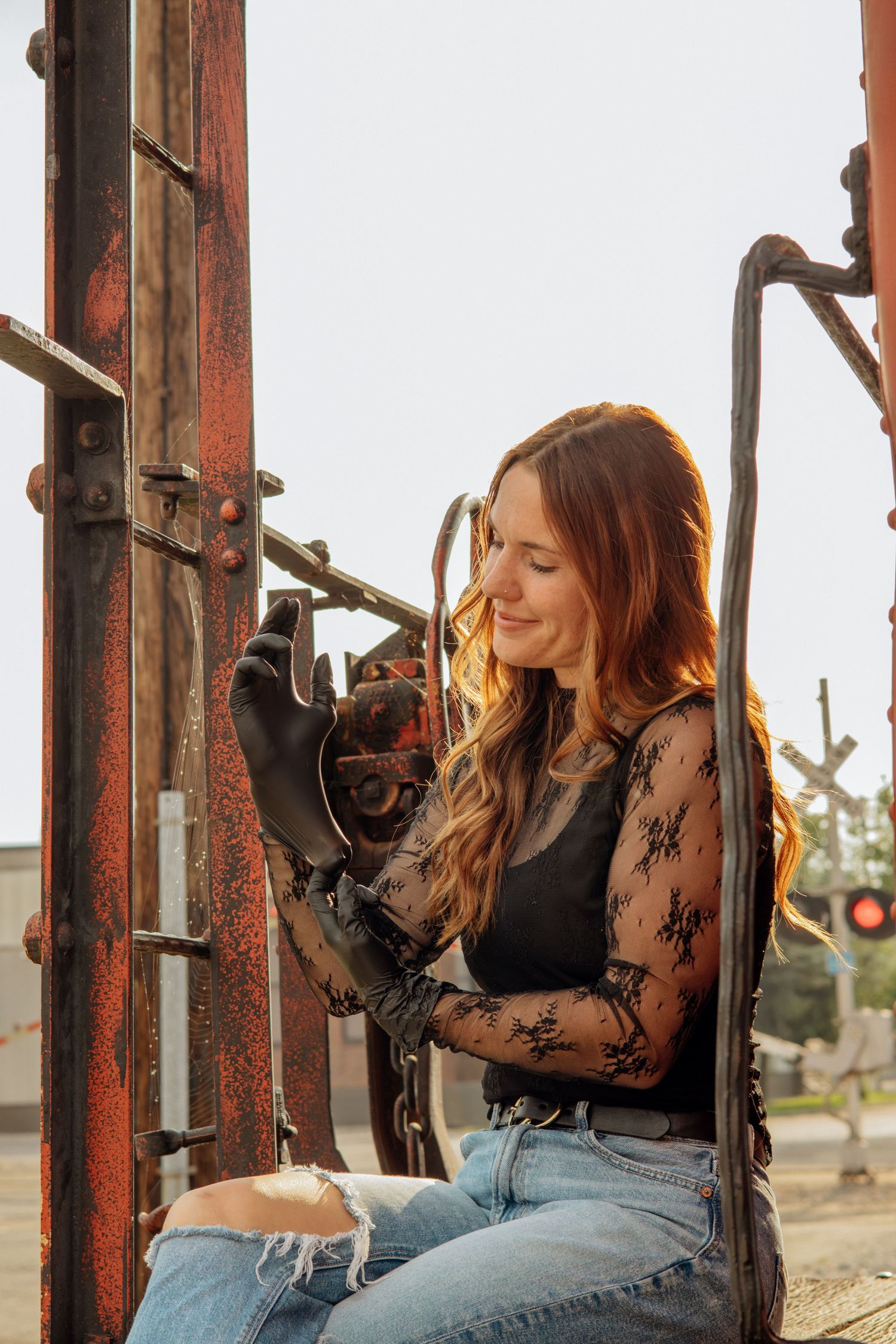 Young woman with long hair wearing lace sleeves and black gloves sits by rusty industrial metal structure at golden hour.