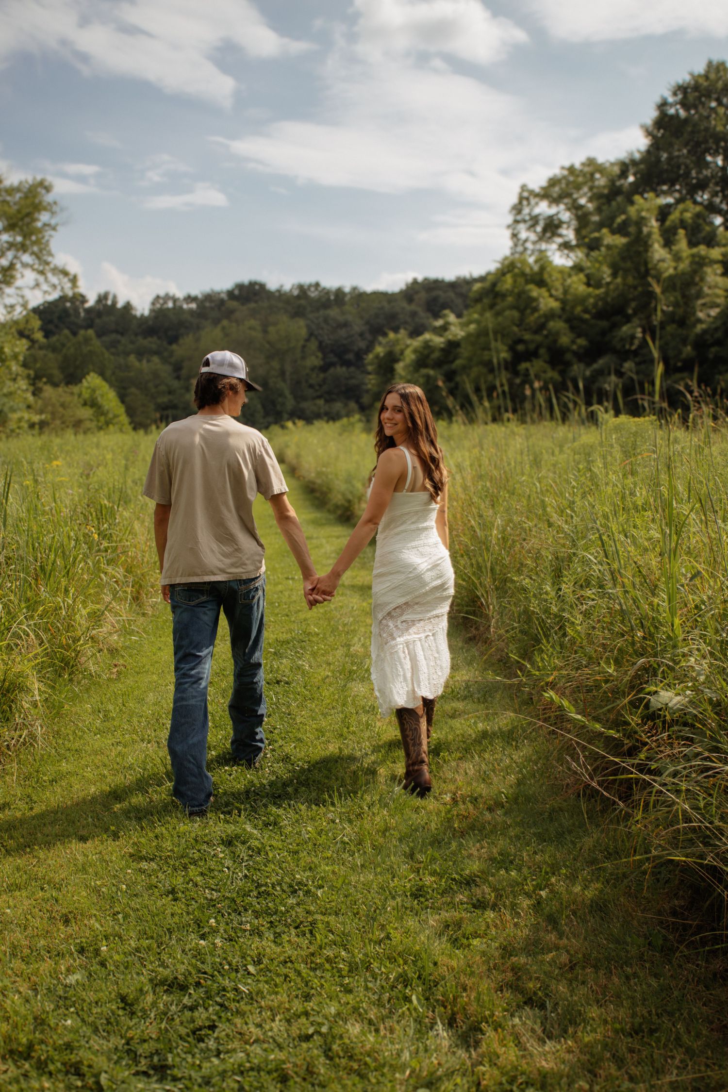 A couple walks hand-in-hand through a lush green field path, woman in white dress, man in jeans and cap.