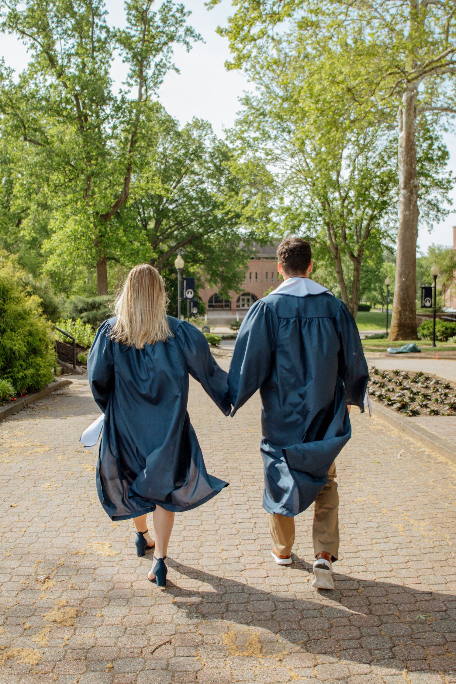 Two graduates in navy caps and gowns walk hand-in-hand along a sunny campus pathway.